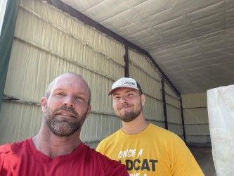 Two men standing in a metal-roofed shed, one in a red shirt and one in a yellow shirt, smiling at the camera.