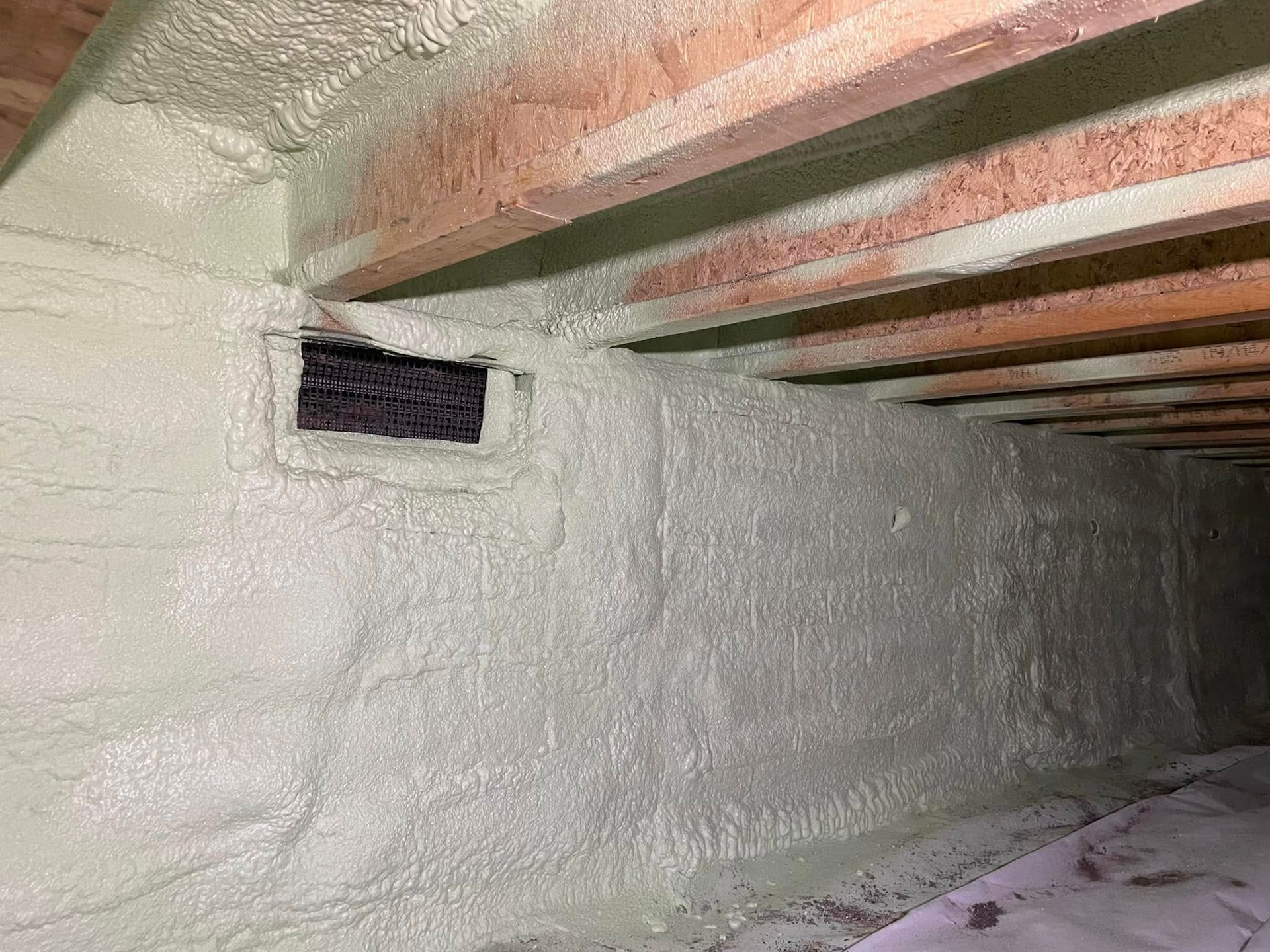 Spray-foam insulated basement wall with exposed wooden ceiling joists and a small vent opening