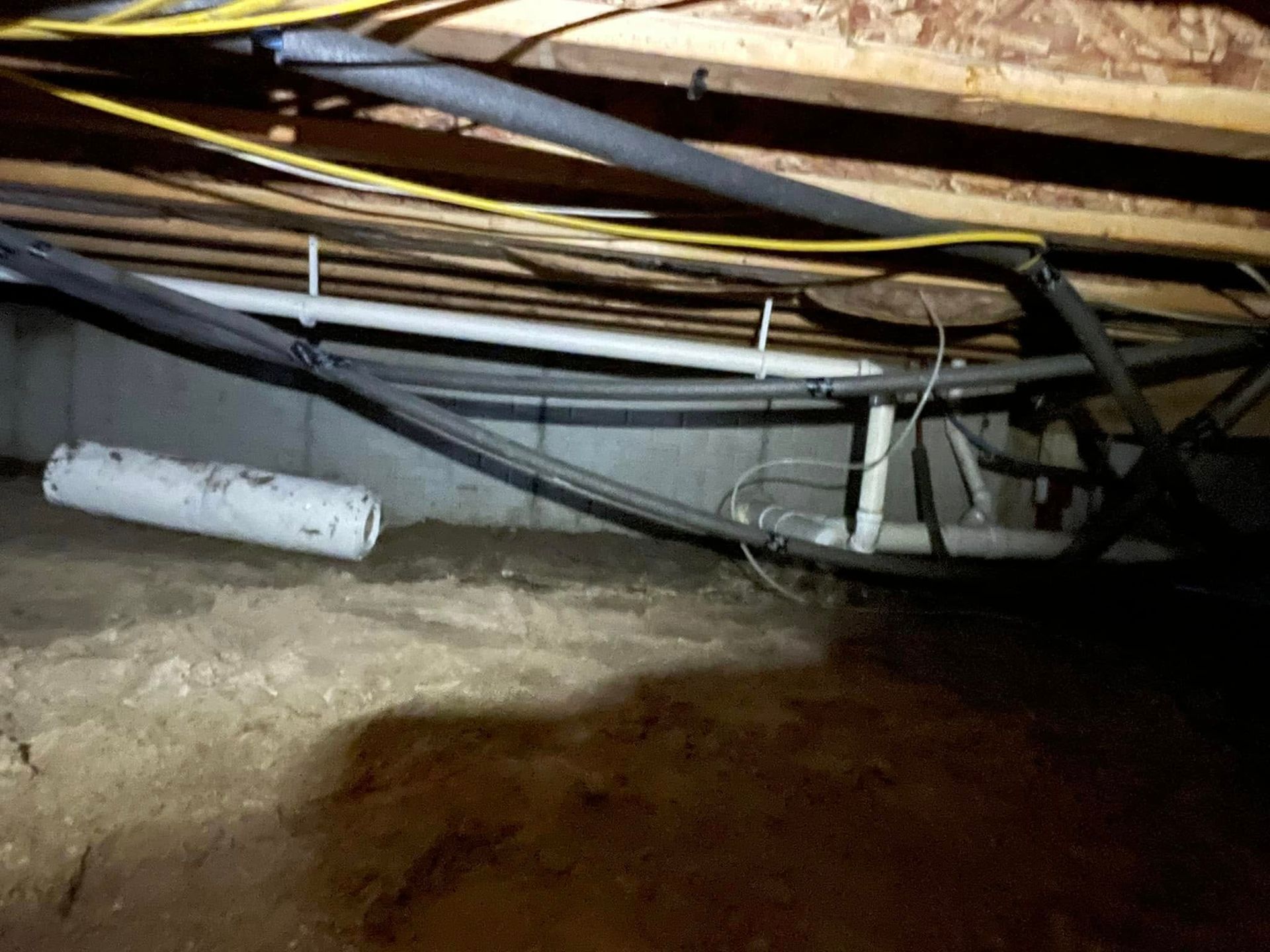 Crawlspace with exposed pipes, cables, and a white cylindrical vent on the dirt floor.