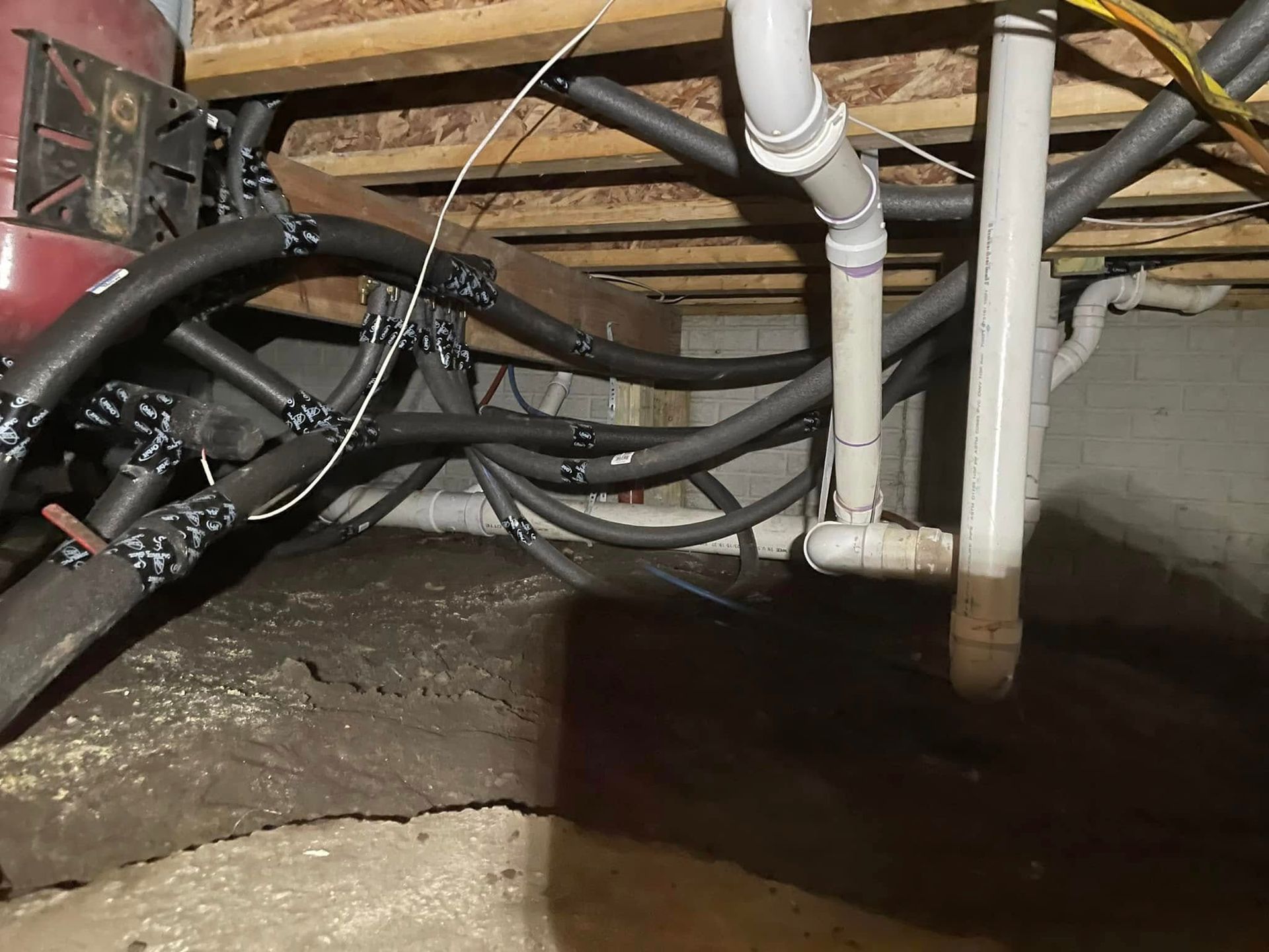 Crawlspace with exposed pipes, wires, and joists above a dirt floor and concrete wall.