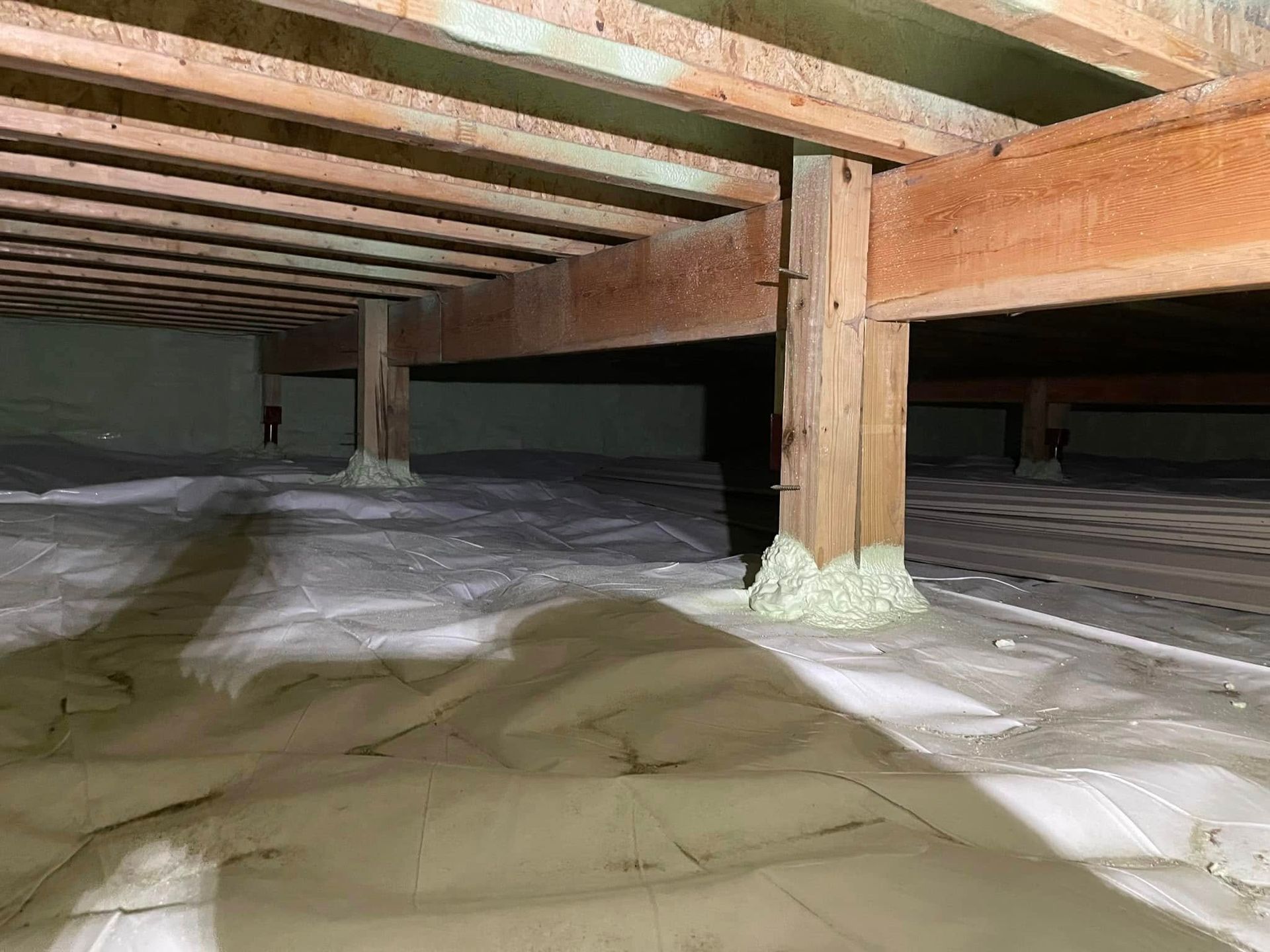 Crawl space with wooden floor joists and support posts above insulation-covered ground