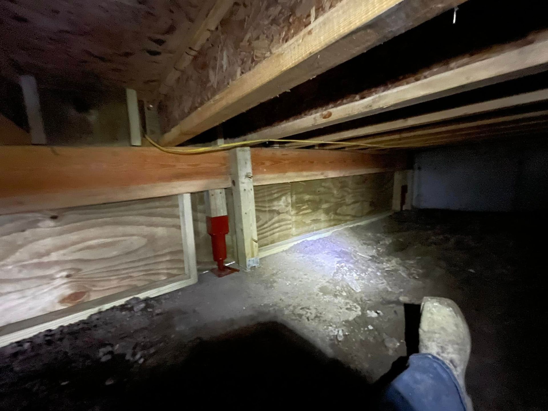 Low crawlspace under wooden floor beams with dirt ground, a red pipe, and a flashlight beam illuminating the area