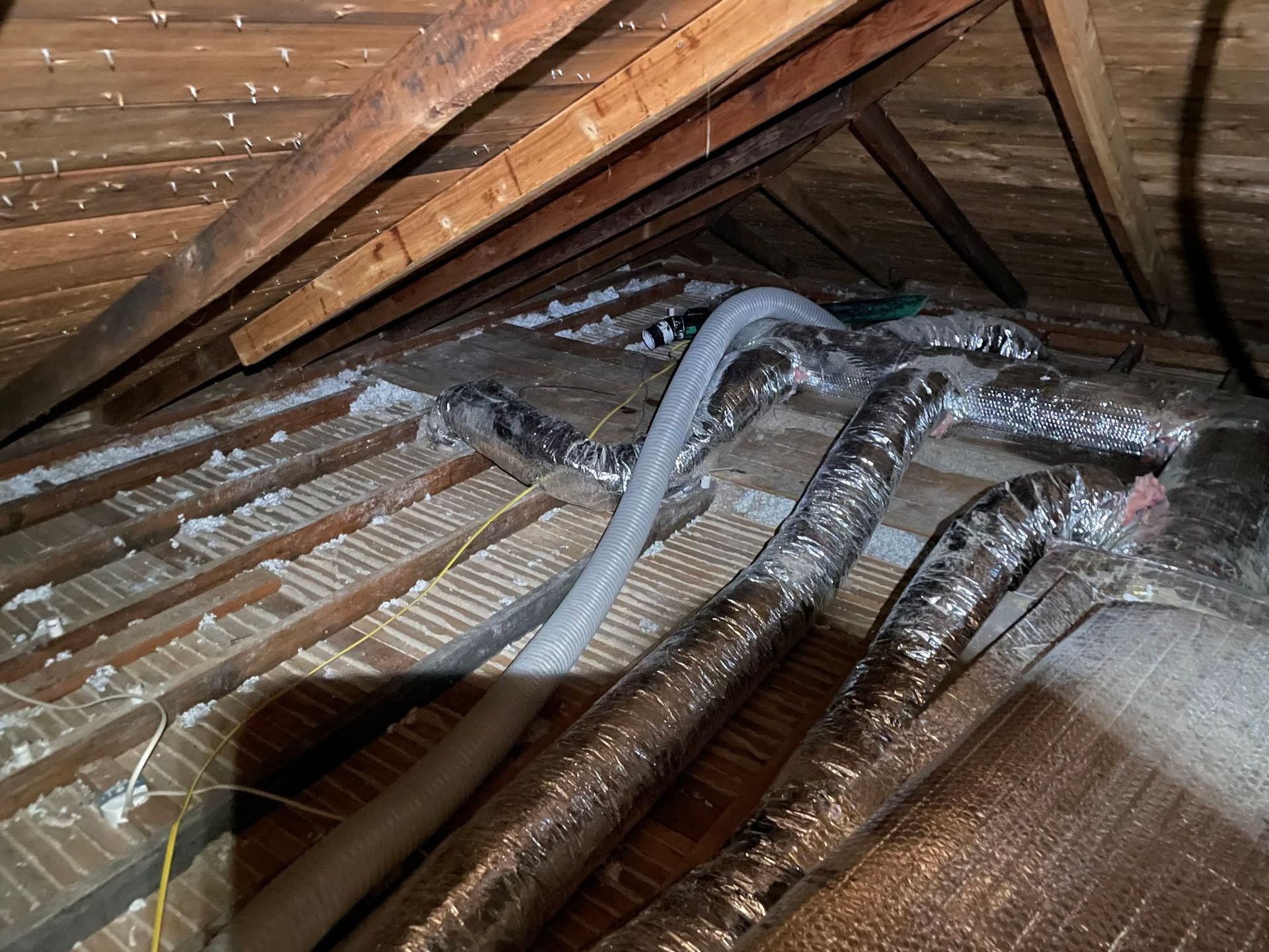 Attic insulation and ventilation ducts under a sloped wooden roof, with exposed rafters and wiring