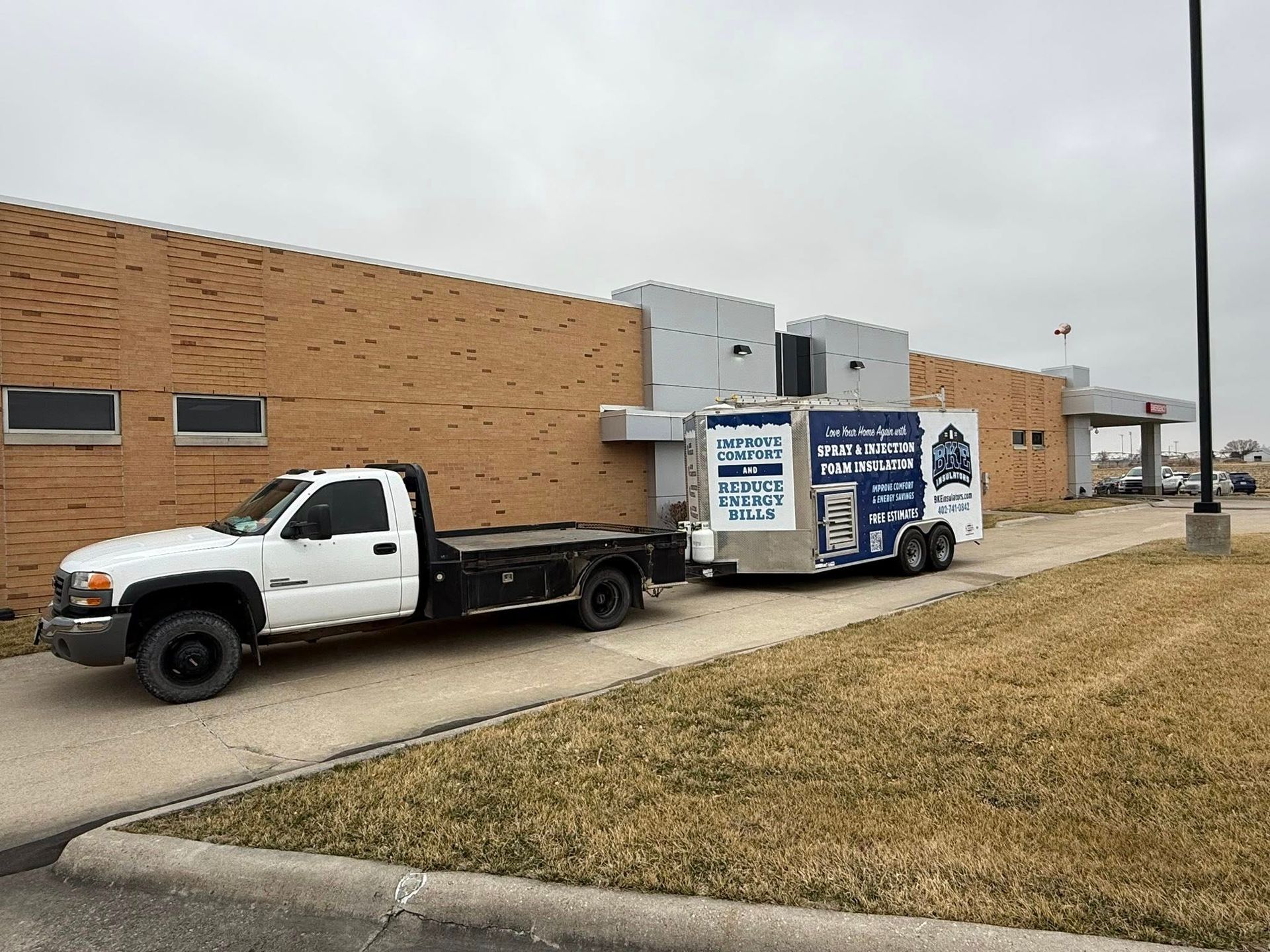 White flatbed truck parked beside a brick building, towing a trailer with a blue-and-white sign.