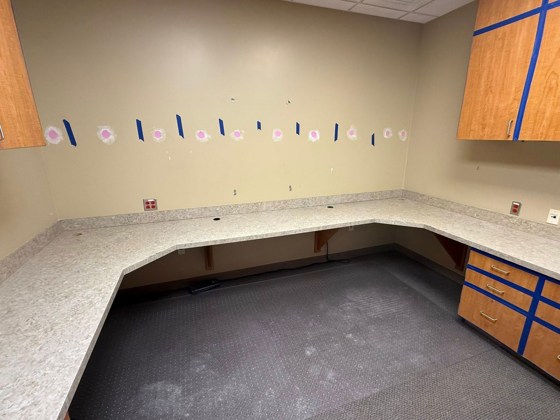 Empty office with beige walls, speckled L-shaped countertop, and gray carpet, showing blue cabinets on the right.