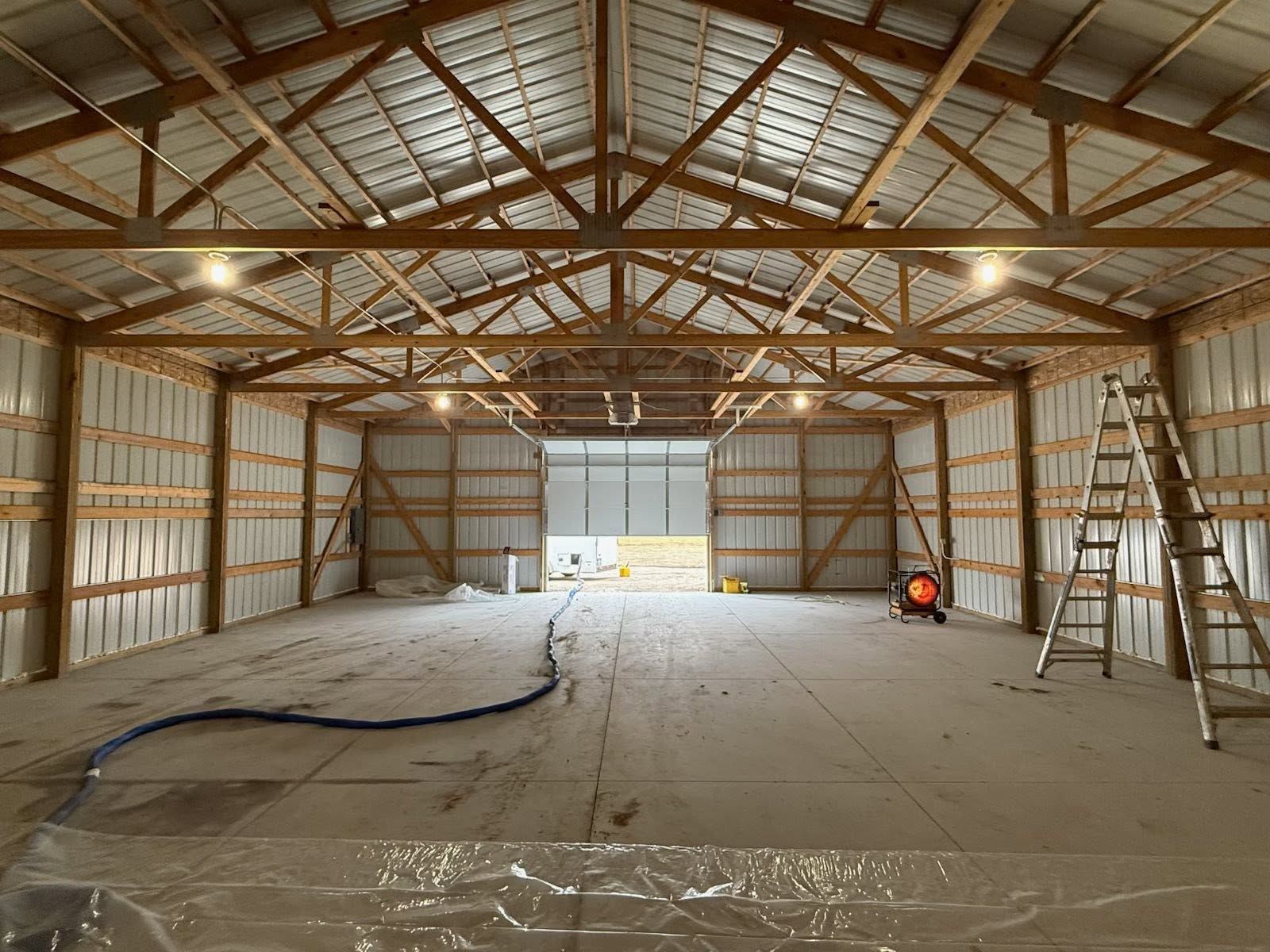 Empty barn interior with exposed wooden beams, concrete floor, ladder, and tools near a large open door