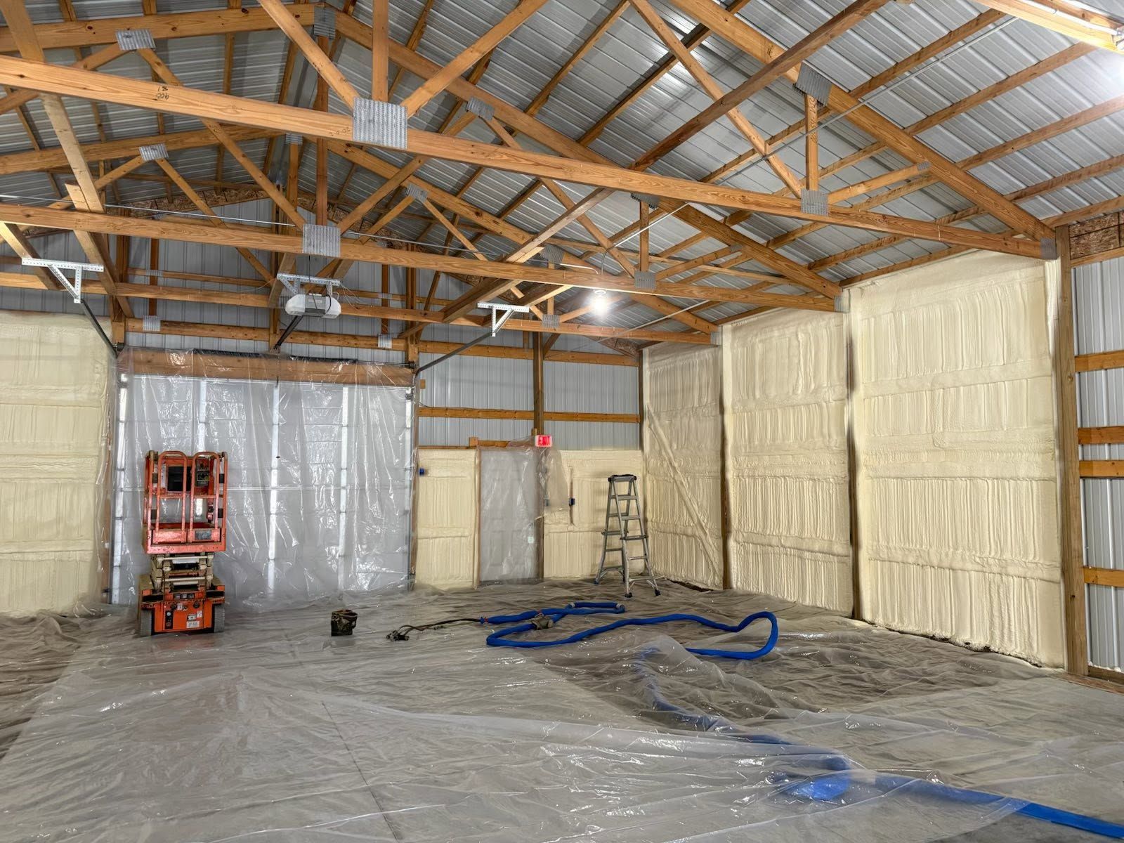 Insulated warehouse interior with exposed roof trusses, plastic-covered walls, and construction equipment on the floor