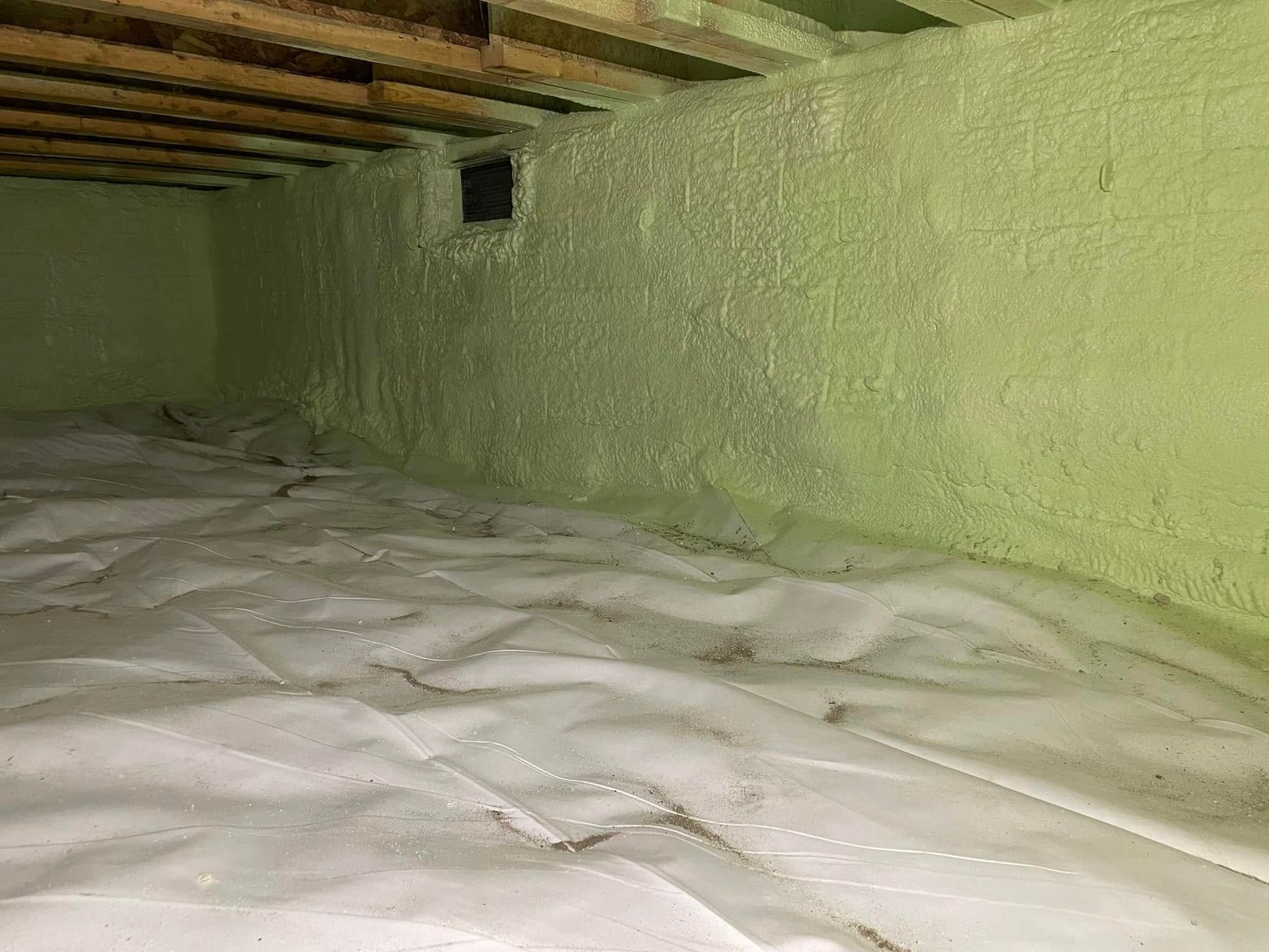 Foam insulation and a white vapor barrier in a low crawlspace with exposed wooden beams