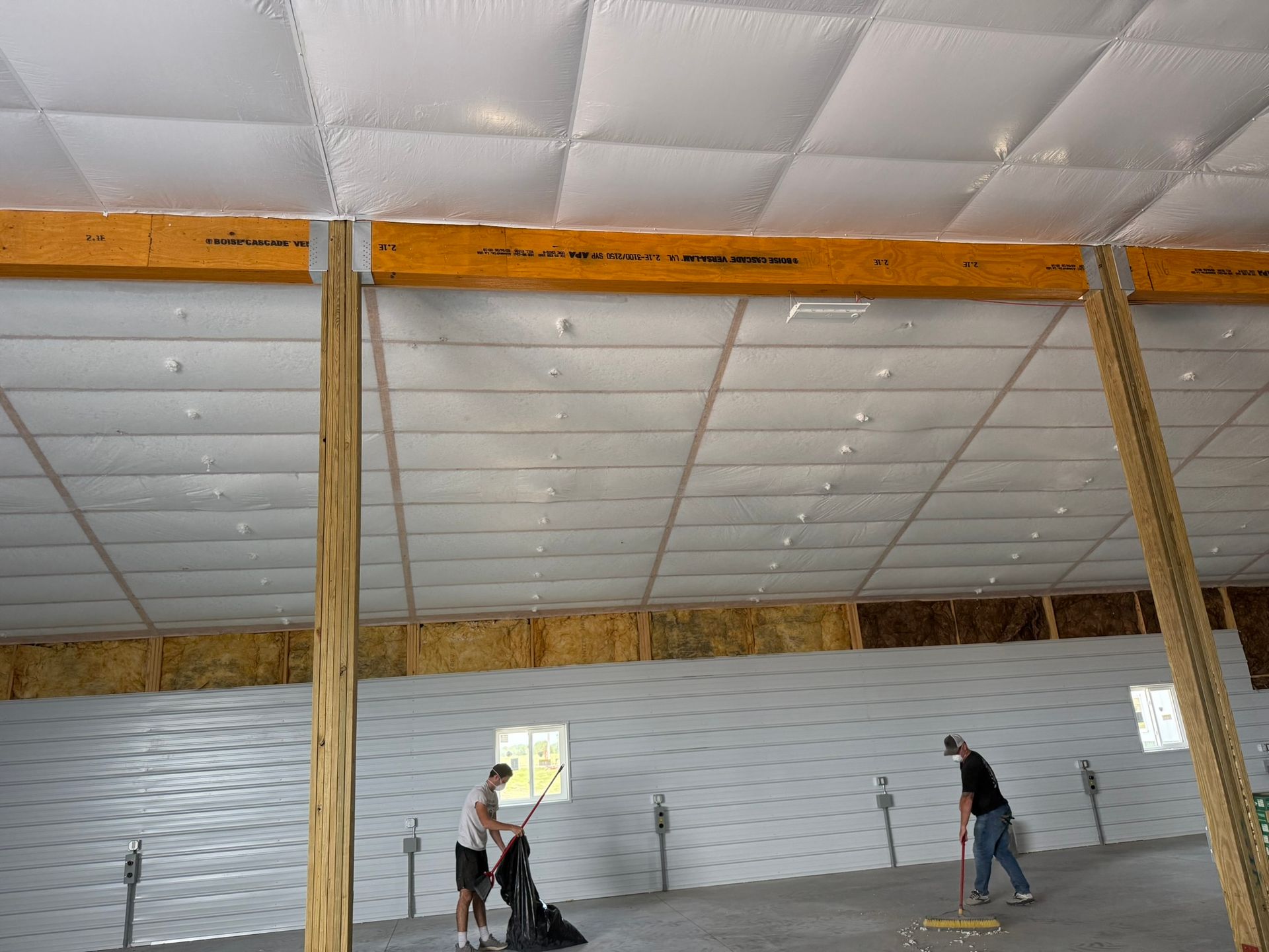 Large warehouse interior with exposed wooden beams and two workers on the floor near the back wall.