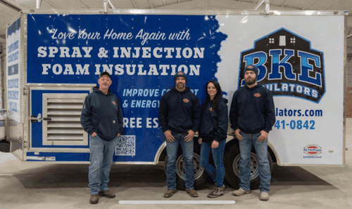 Four people standing in front of a BKE spray foam insulation trailer with blue-and-white branding.
