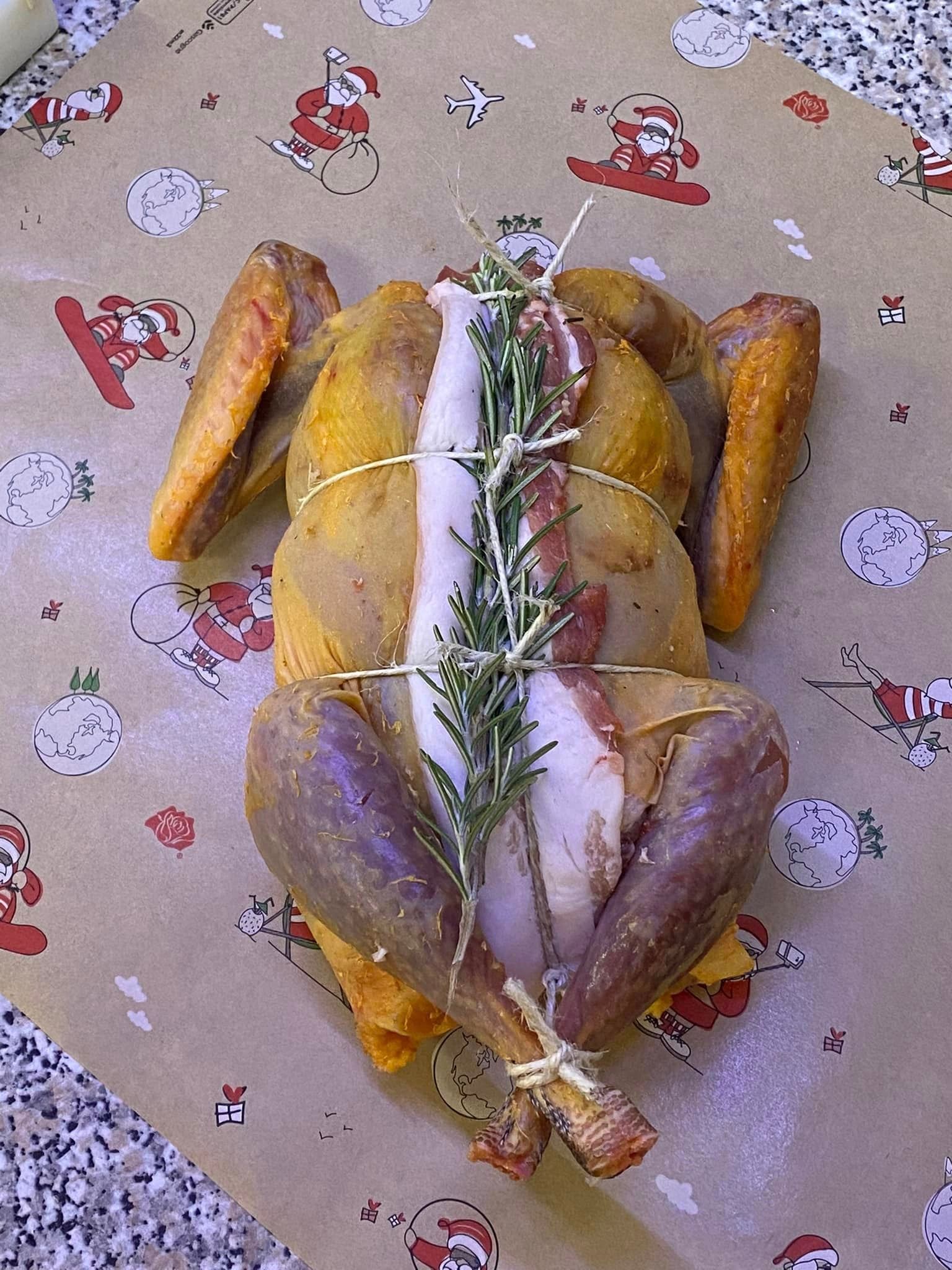 Un pollo intero crudo e condito, legato con spago, guarnito con strisce di pancetta e rosmarino fresco, il tutto avvolto in carta da regalo festiva.