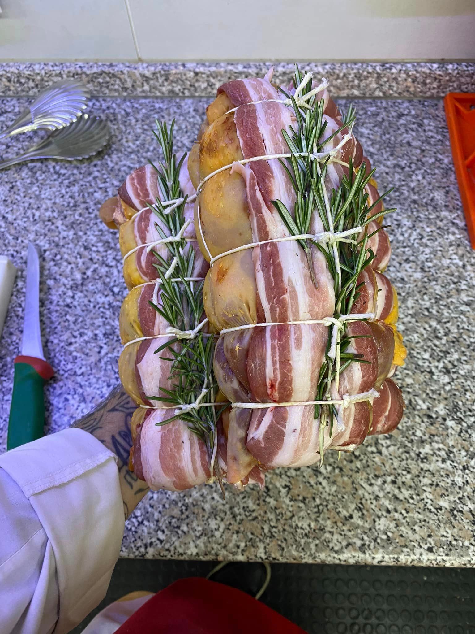 Due carcasse di pollo avvolte in pancetta e rosmarino con spago da cucina, tenute sopra un bancone di granito grigio.