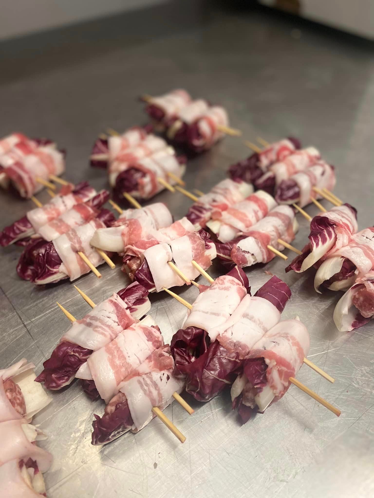 Piccoli fagottini di carne cruda avvolti in strisce di pancetta e fissati con spiedini di legno, disposti su una superficie in acciaio inossidabile.
