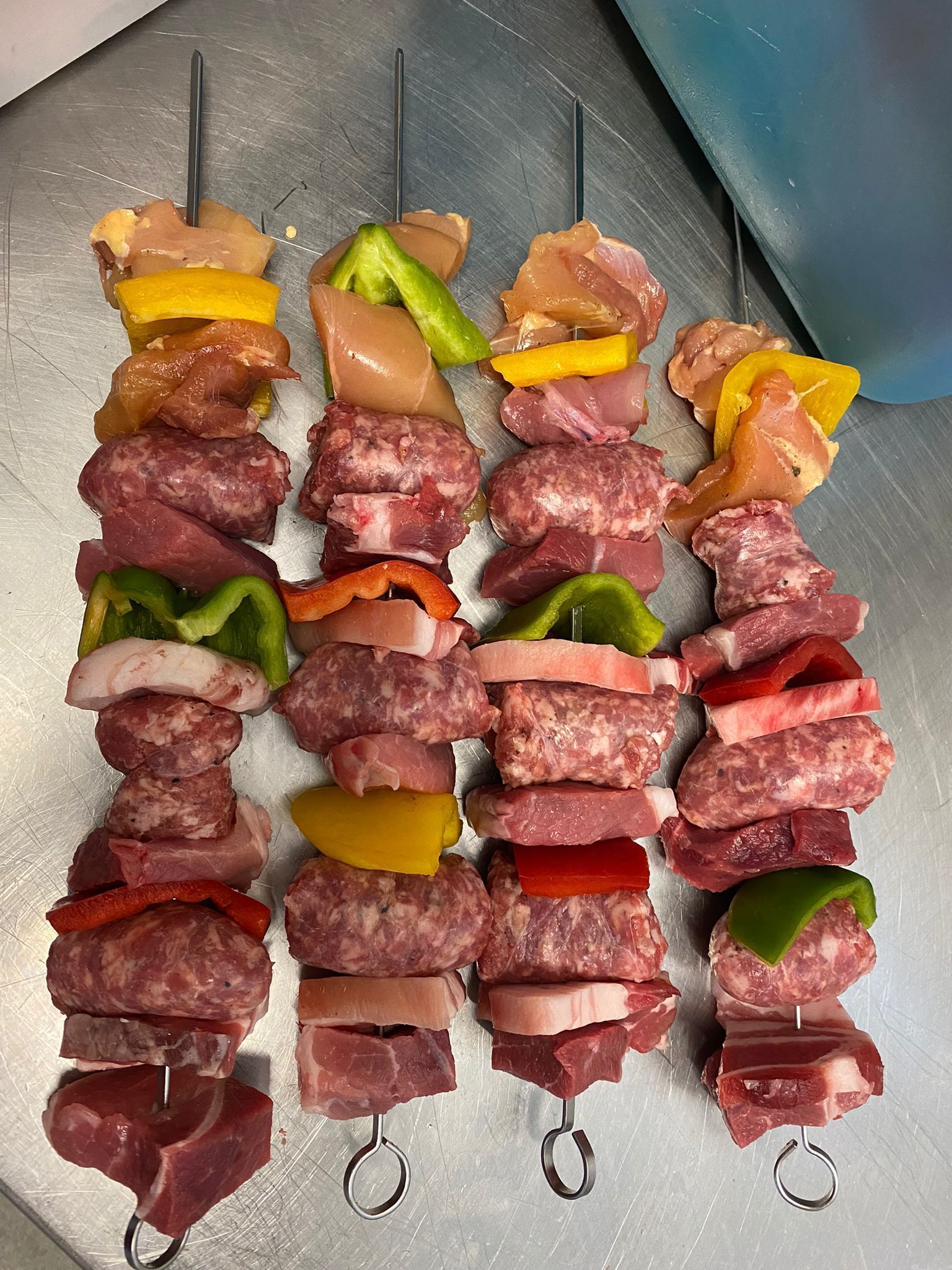Quattro spiedini di carne cruda e verdure disposti uno accanto all'altro su una superficie metallica.