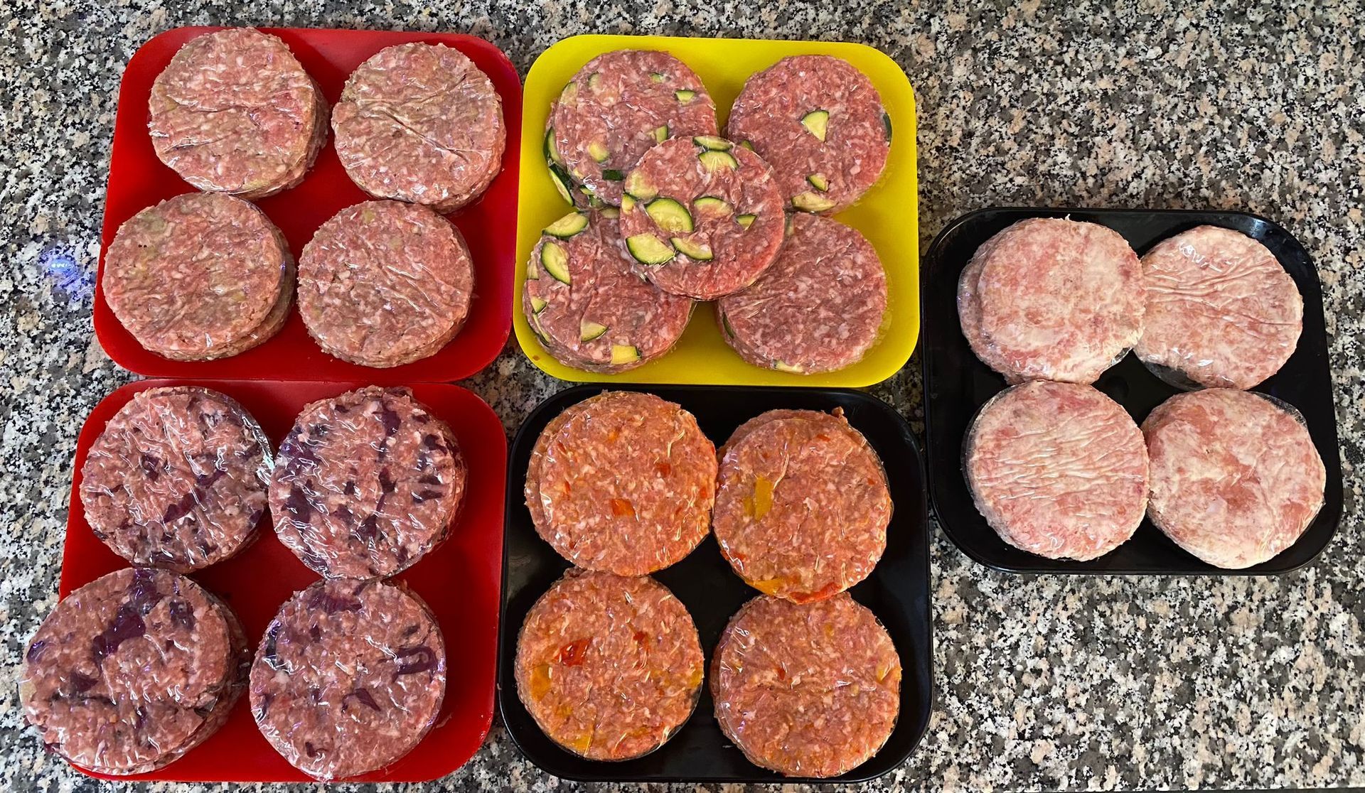 Su un piano di lavoro maculato, cinque piatti quadrati colorati contengono vari tipi di hamburger crudi.
