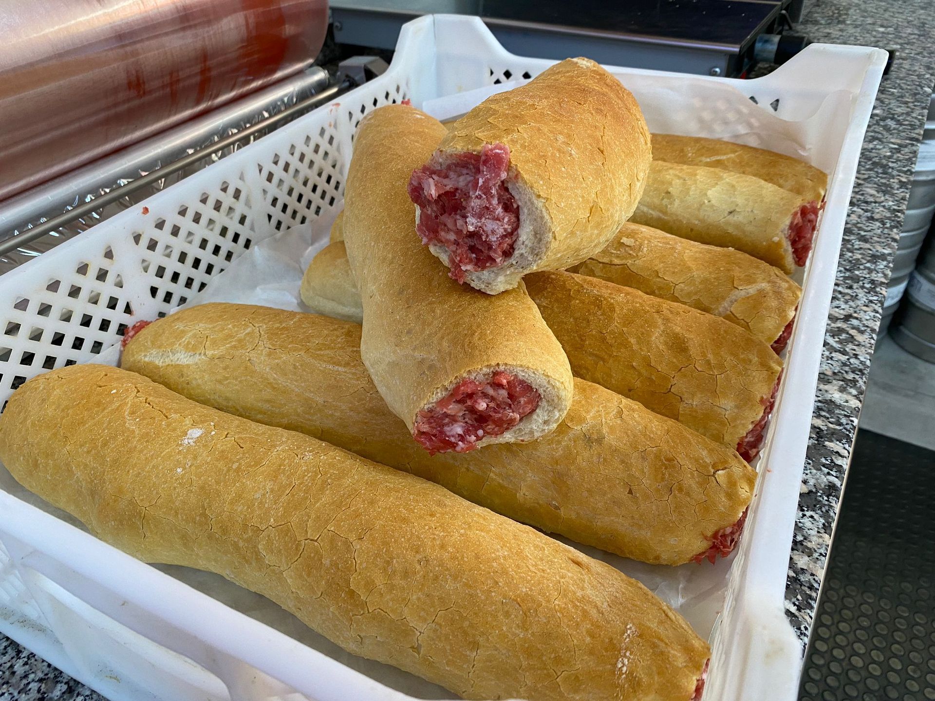 Un vassoio di plastica bianca pieno di lunghe salsicce avvolte nel pane, una delle quali tagliata per rivelare il ripieno di carne cruda.