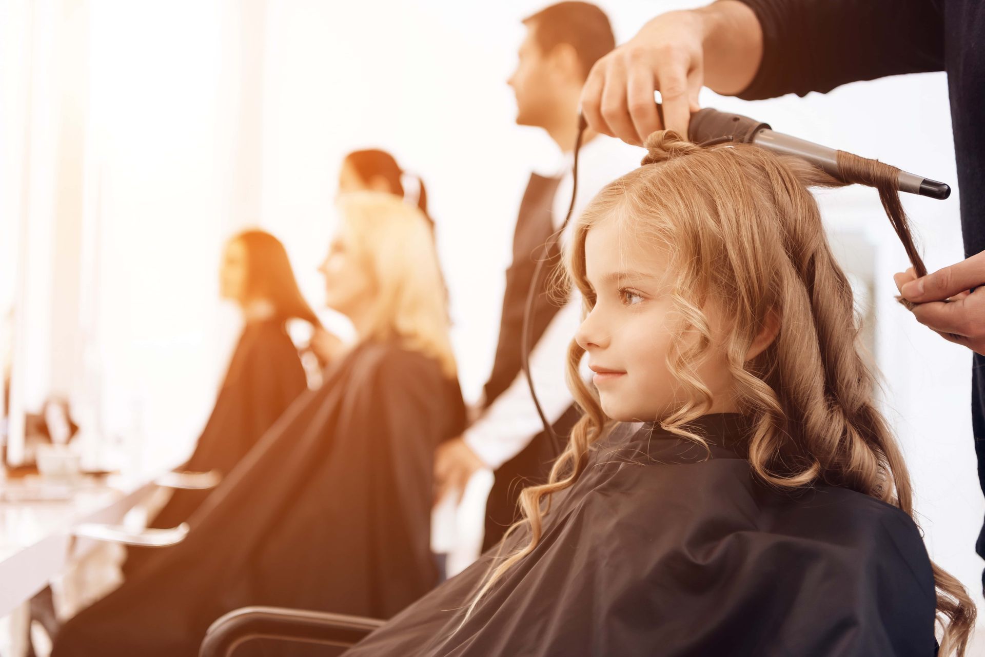 Girl getting her hair curled with a curling iron at a salon. Other clients and stylists in the background.