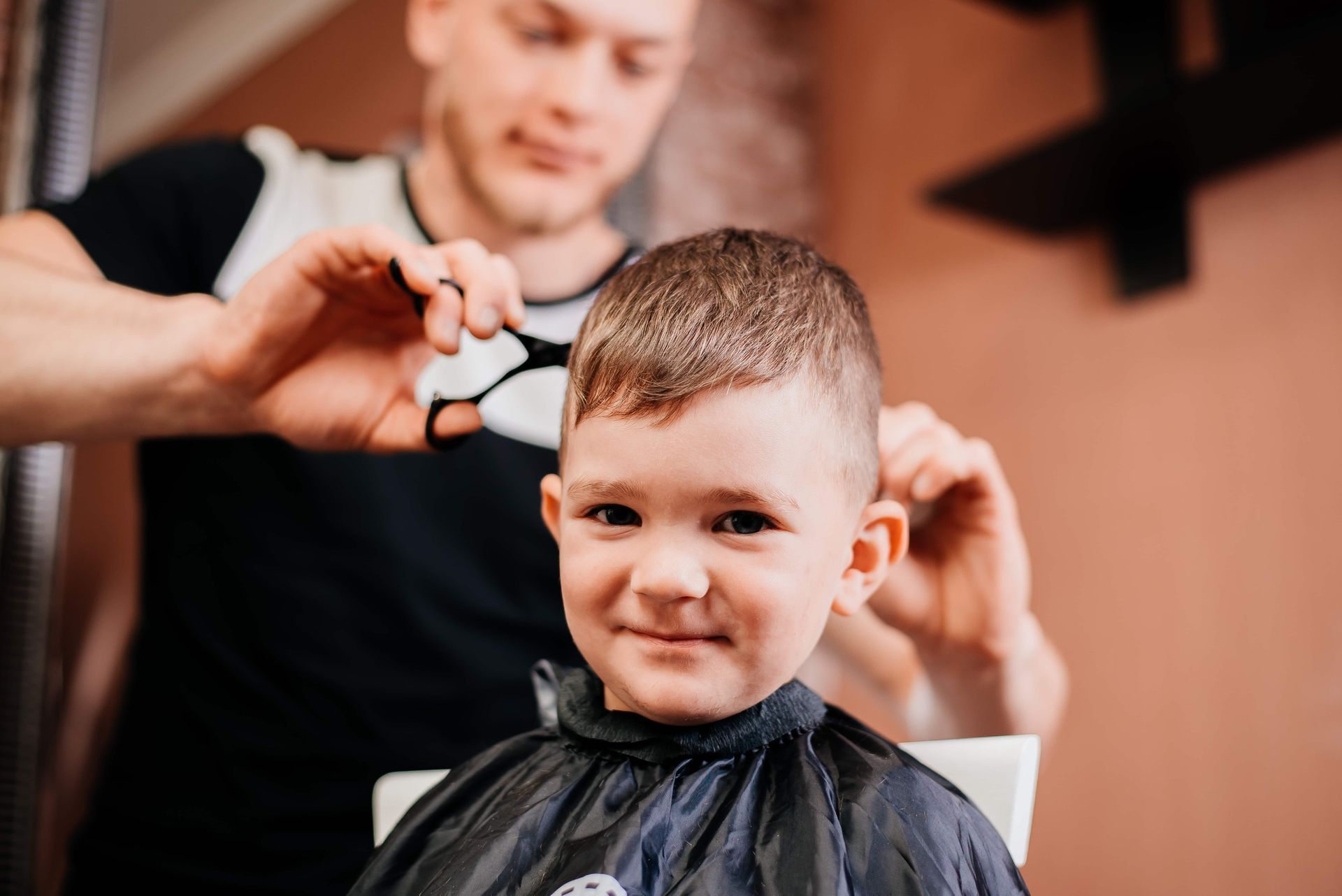 Boy getting a haircut at a salon. The boy smiles while the barber cuts his hair with scissors.
