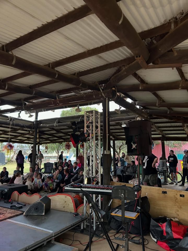 People Seated Under a Covered Pavilion Near a Stage with Speakers and Audio Equipment — Blatherskite Park in Ilparpa, NT