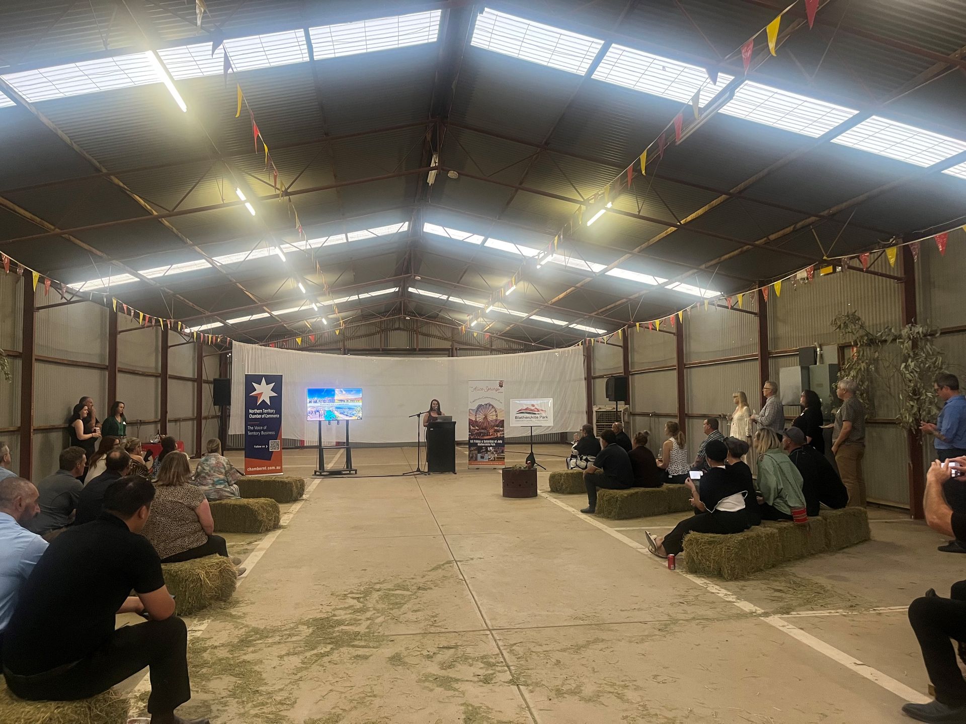 People Seated in a Large Hall Facing a Stage with Banners and Flags During an Indoor Event — Blatherskite Park in Ilparpa, NT