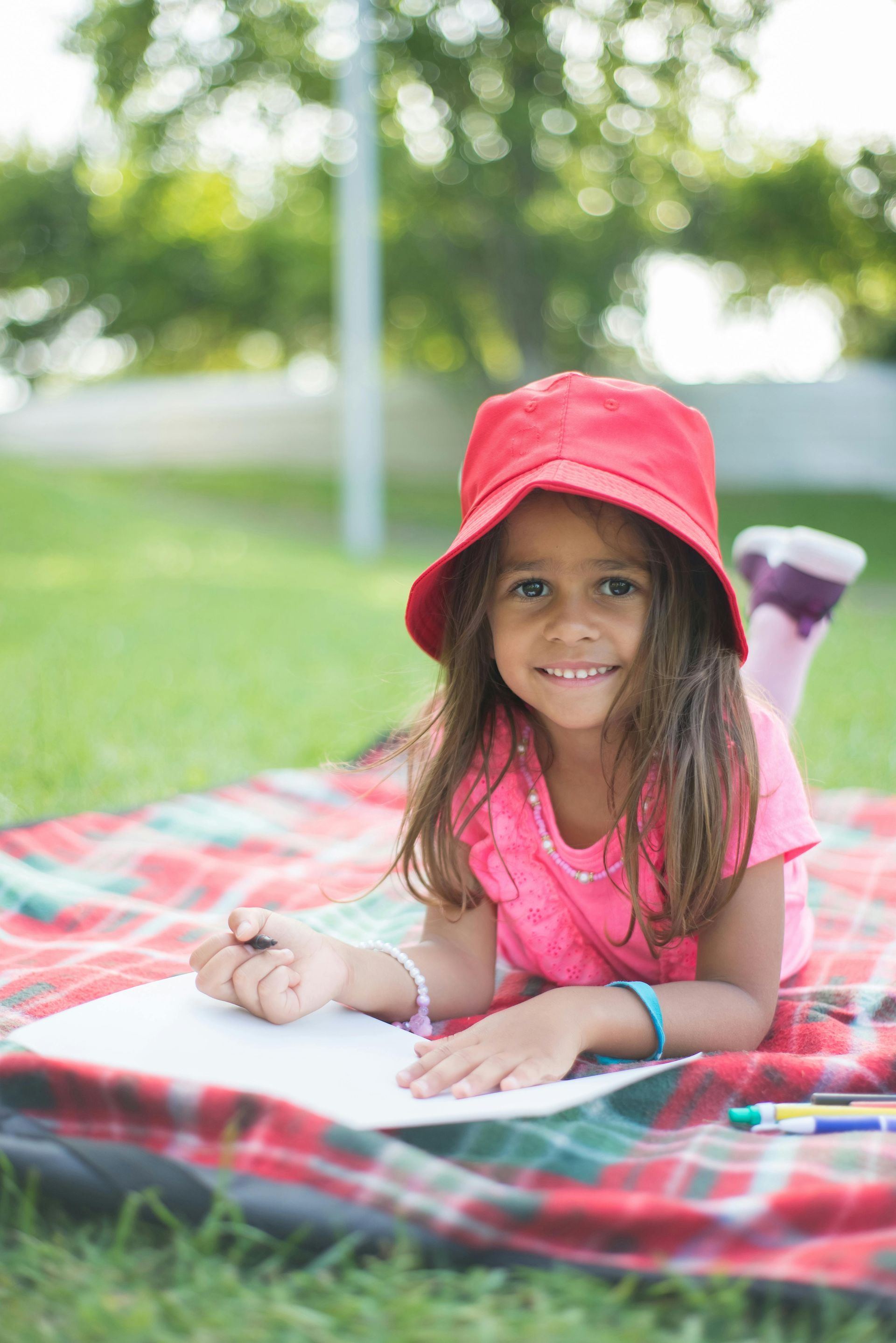 Child in a red sunhat lying on a picnic blanket and smiling outdoors in a grassy park.