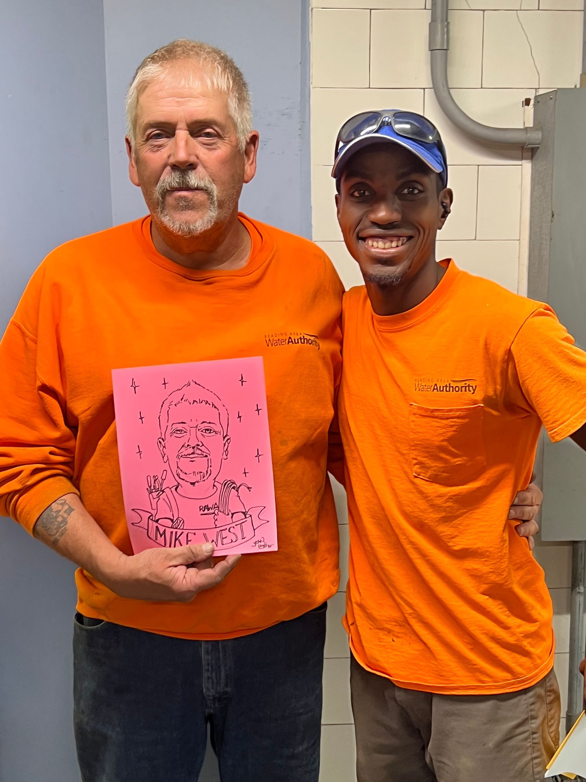 Two men in orange shirts, one holding a pink drawing, smiling. Indoors, against a wall.