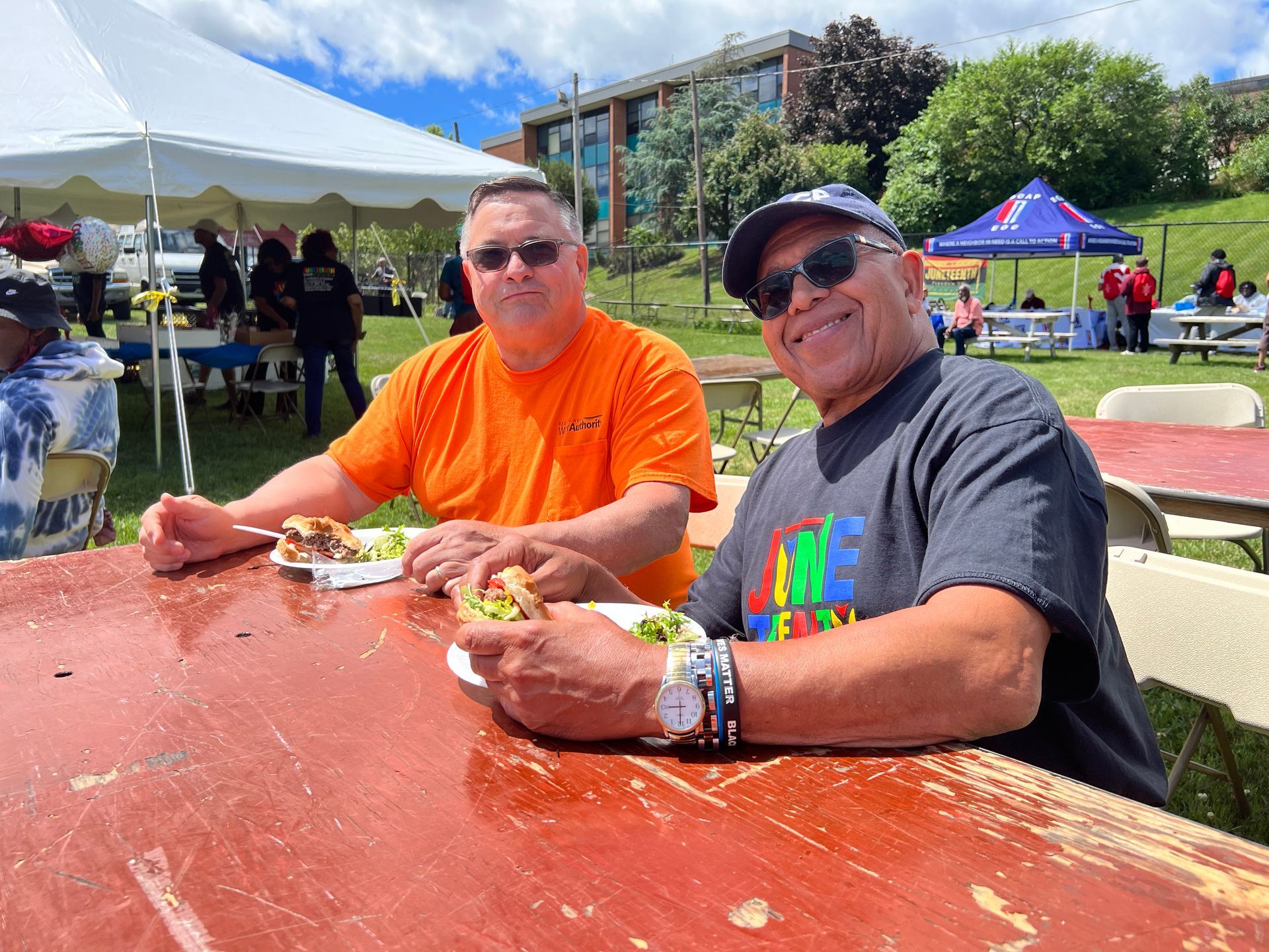 Two men seated at a picnic table, eating hamburgers outdoors. One wears orange, the other a black tee.