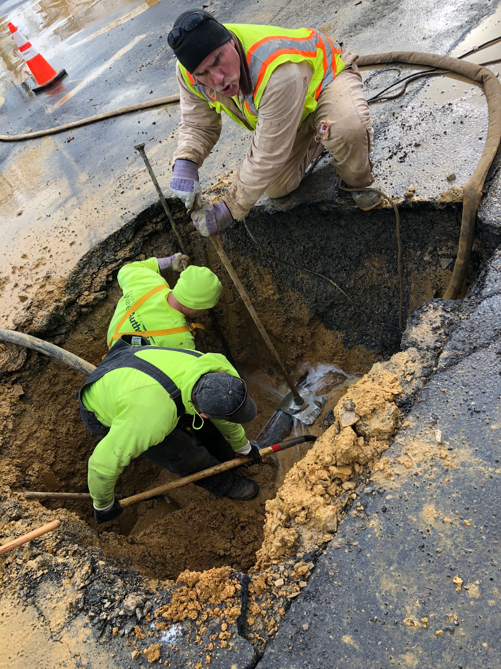 Three workers in safety vests repair a road. One kneels in a hole, two stand above, all using tools.