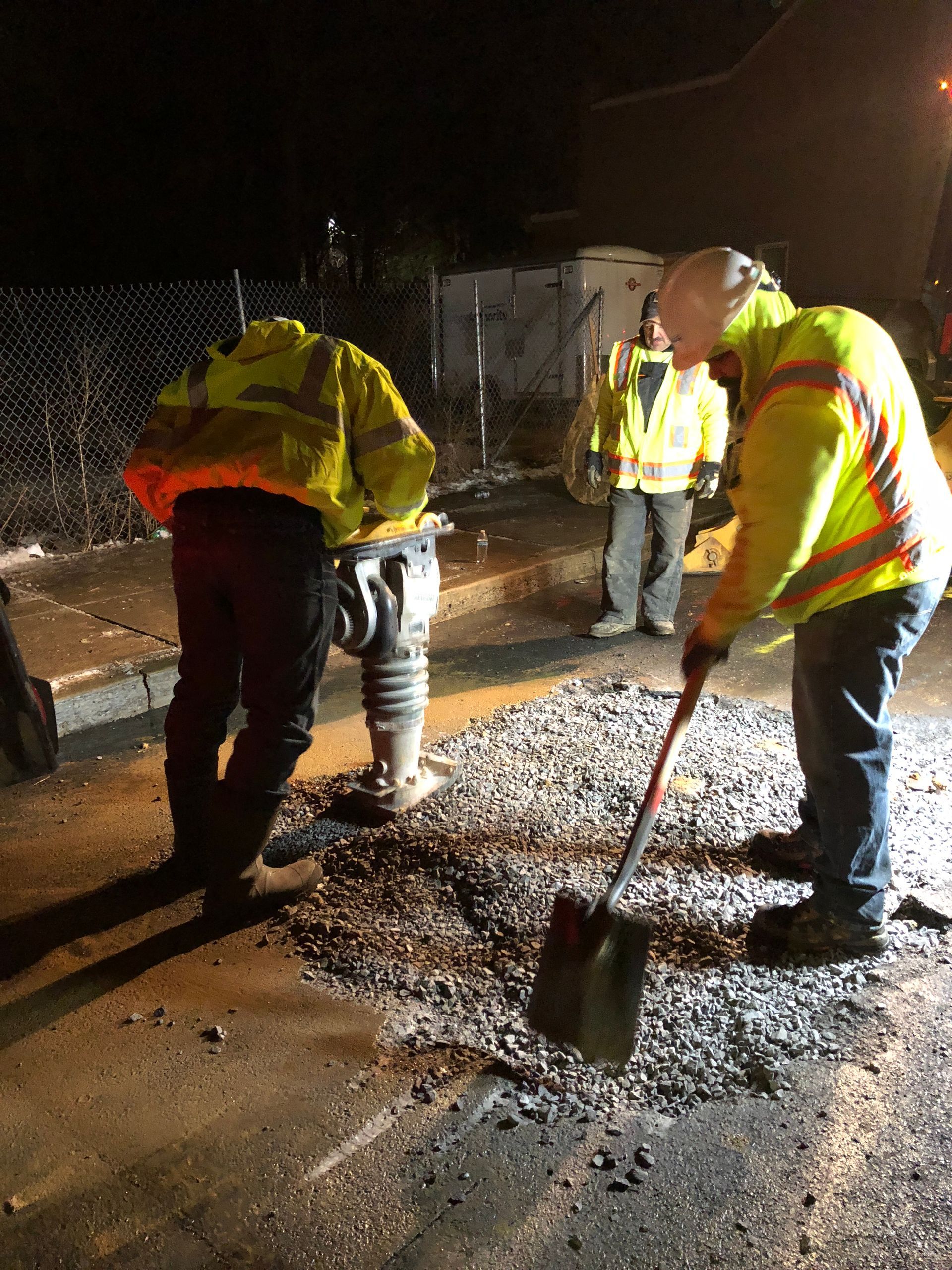 Road workers repairing asphalt at night; one using a tamper, another shoveling gravel, and a third observing.