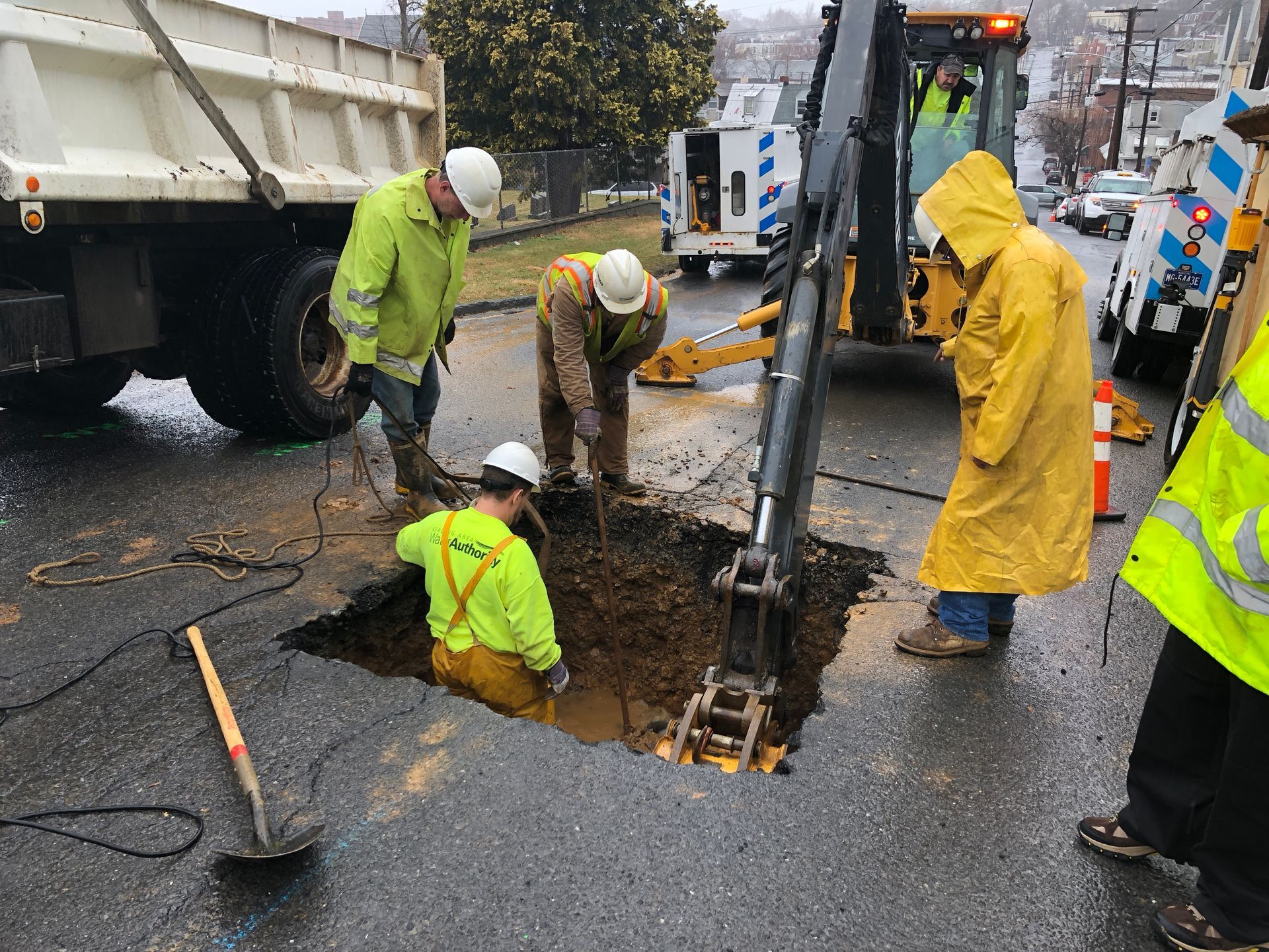 Workers in yellow rain gear and hard hats repair a road, with an excavator, truck, and tools present.