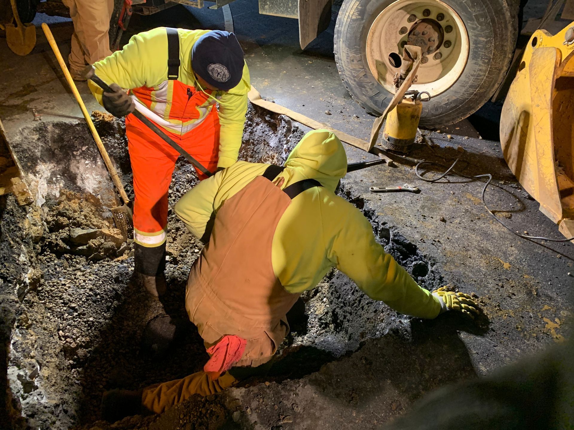 Two workers in yellow and orange safety gear repairing asphalt road.