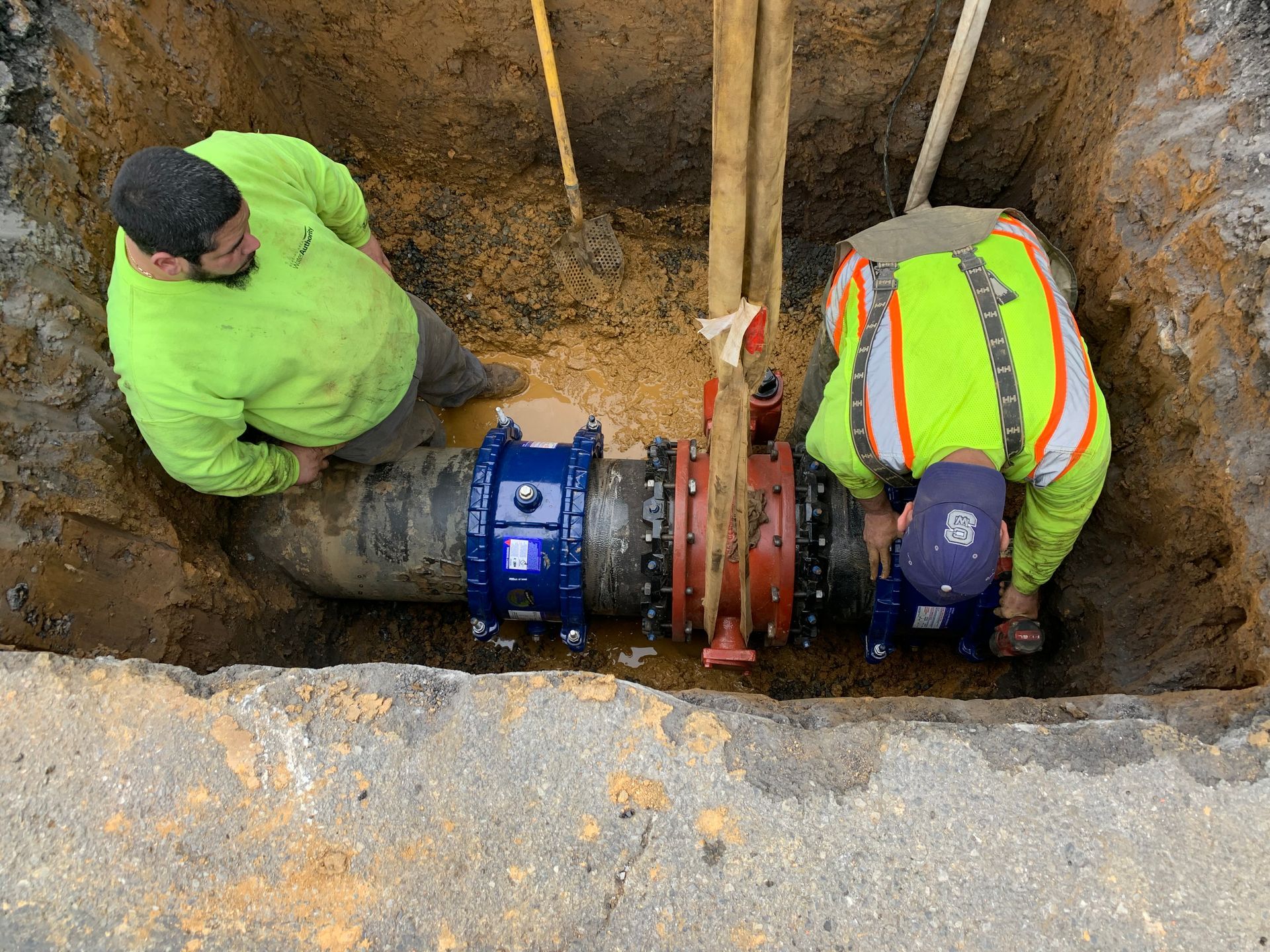 Two workers in a trench connecting water pipes, wearing safety vests and hard hats.