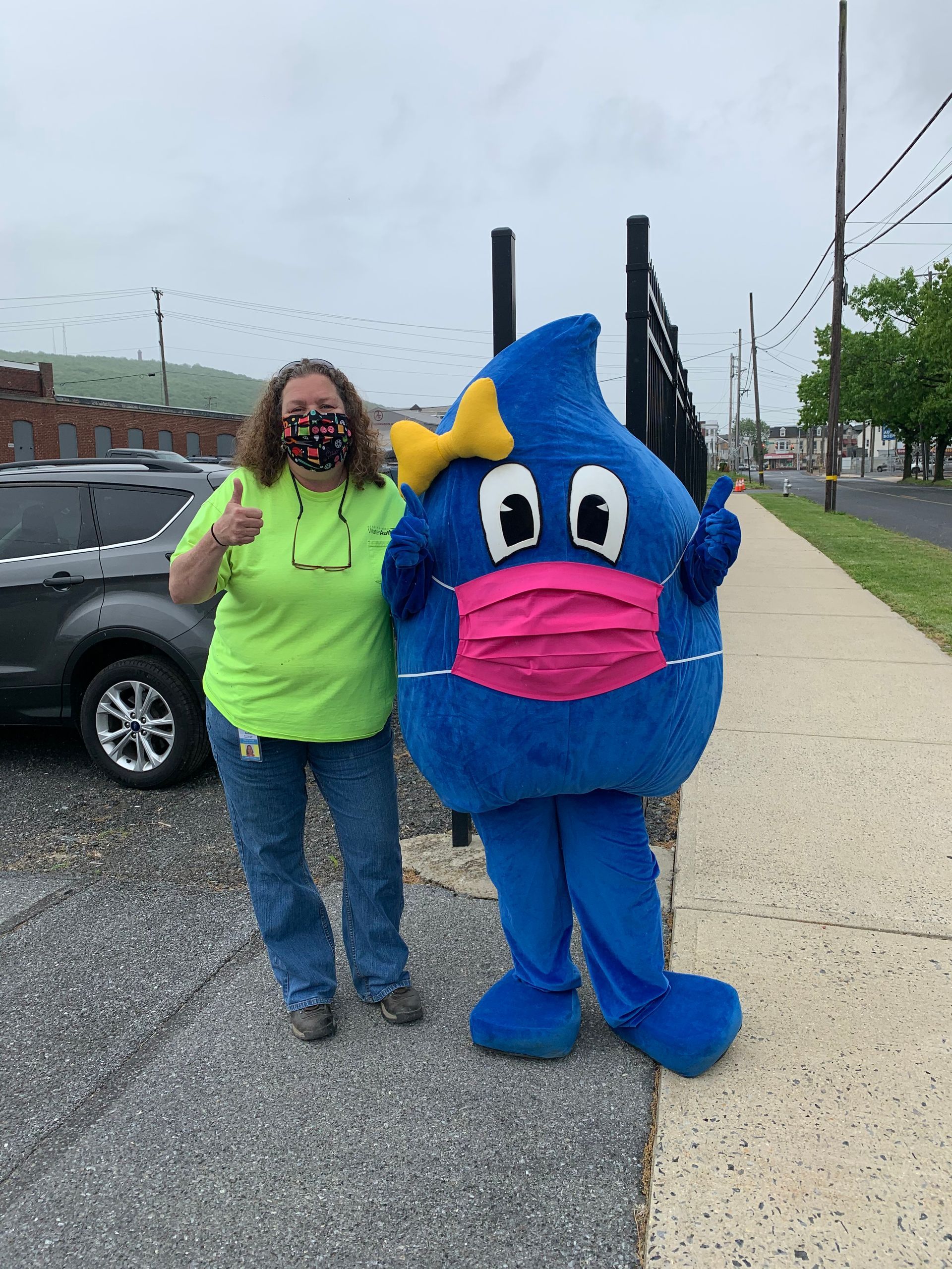 Woman and water drop mascot wearing masks, giving thumbs up, on sidewalk.