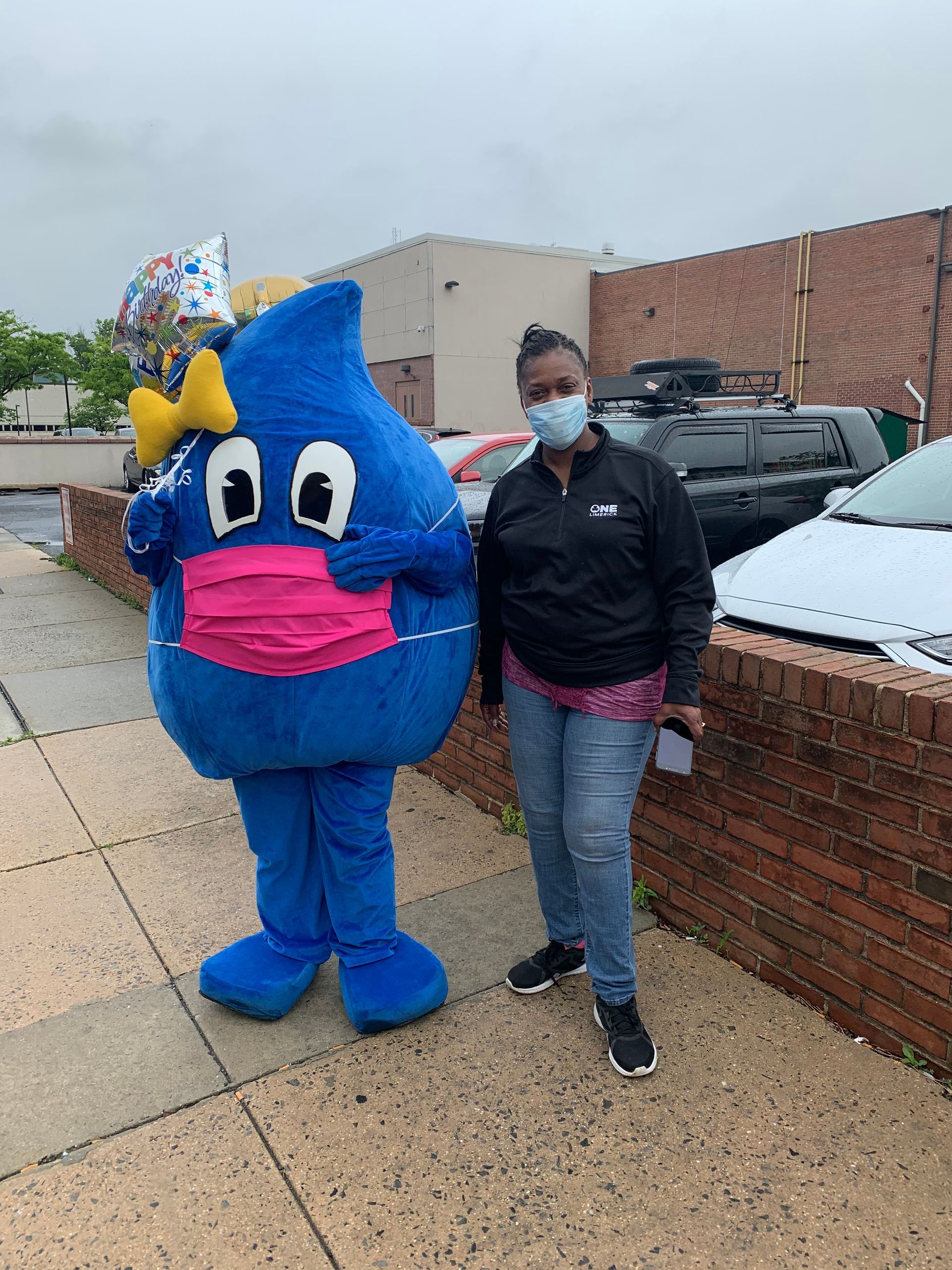 Person and water droplet mascot wearing masks, standing on a sidewalk. Cars in background.