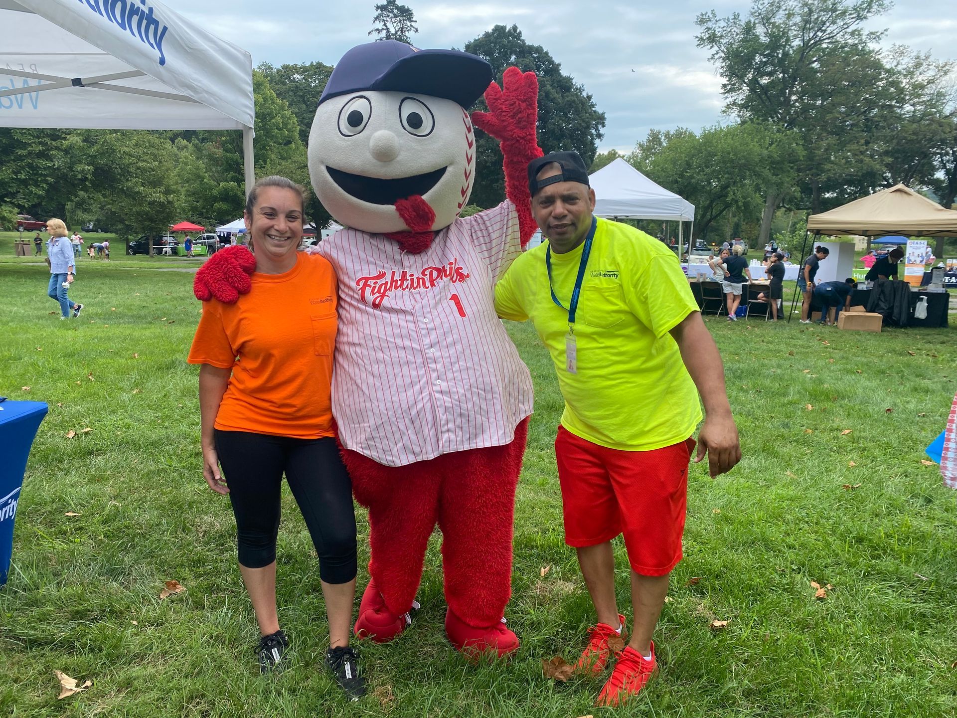 Woman and man pose with a baseball mascot in a park. They are wearing casual clothing.