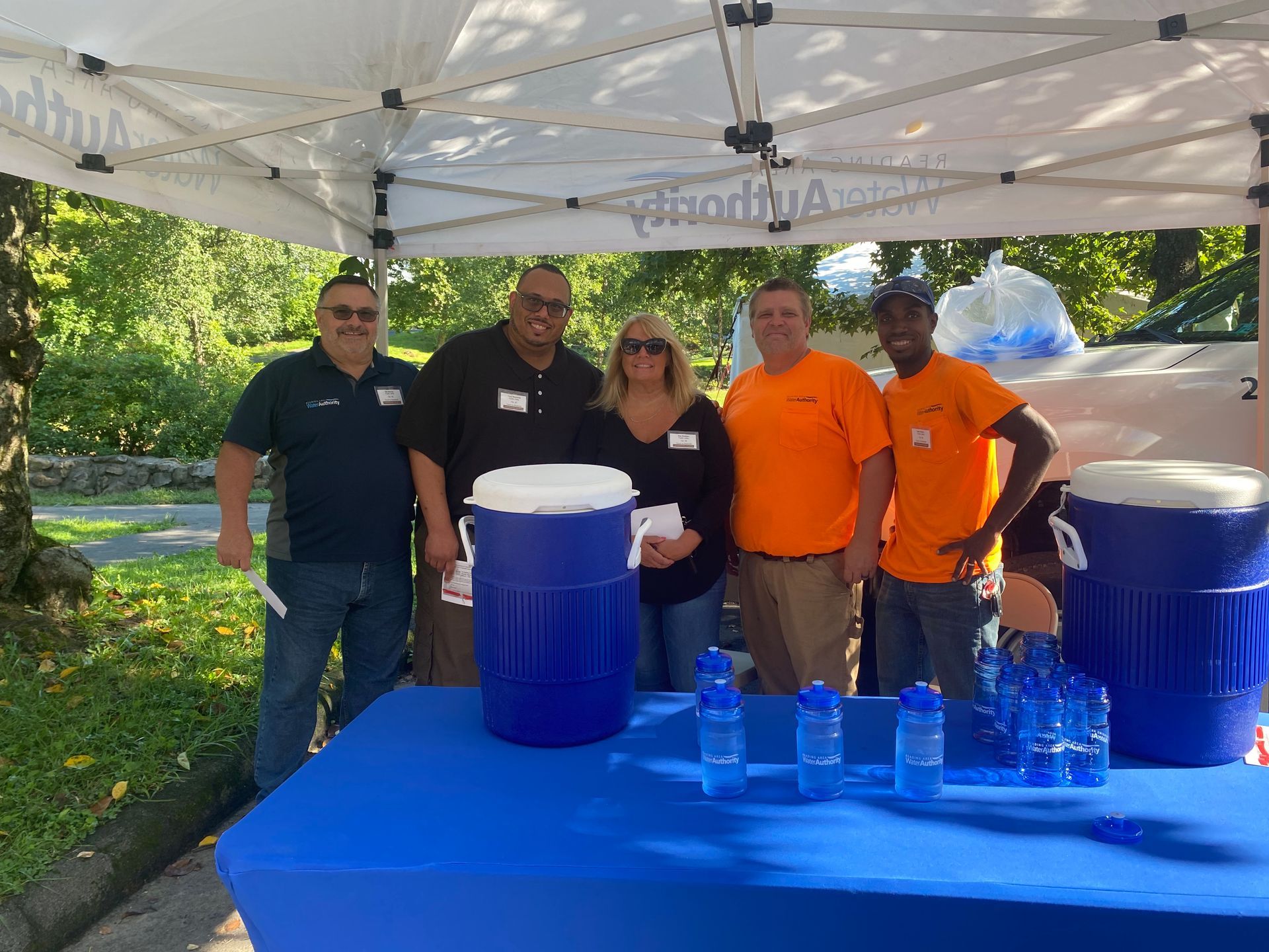 People at a booth with blue table, water cooler, and bottles under a white tent.