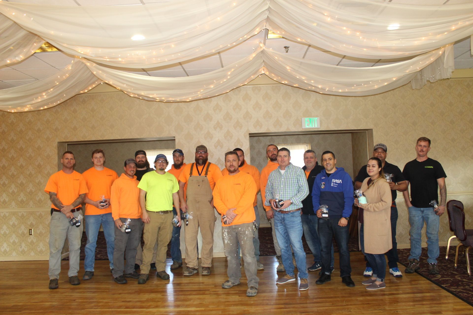 Group of people in bright orange shirts and work attire posing indoors; festive lights and patterned wallpaper.