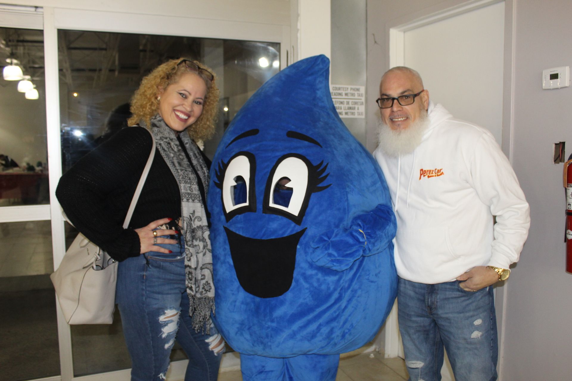Woman and man pose with a blue water droplet mascot, smiling inside a building with a glass door.