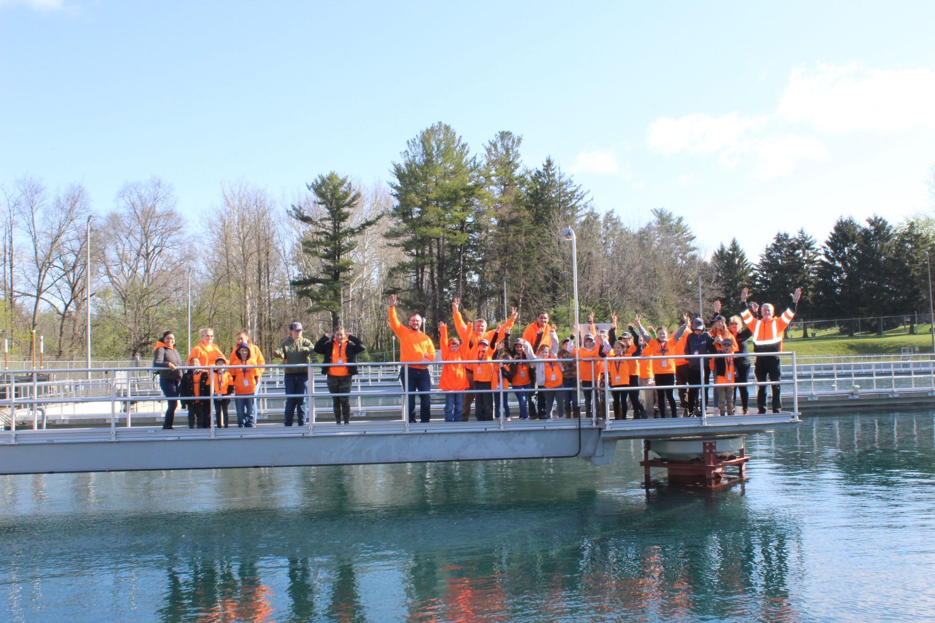 Group of people in orange vests wave from a platform over water. Trees and blue sky in background.