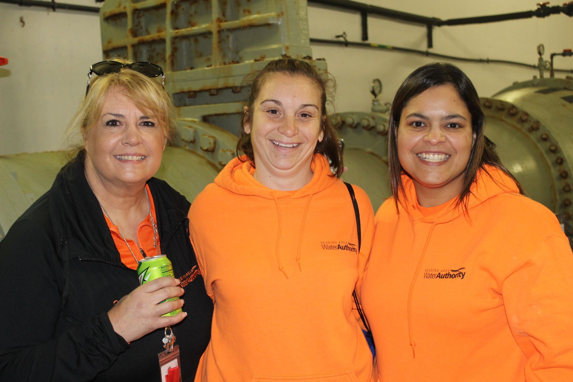 Three women smiling, wearing orange hoodies, standing near industrial equipment.