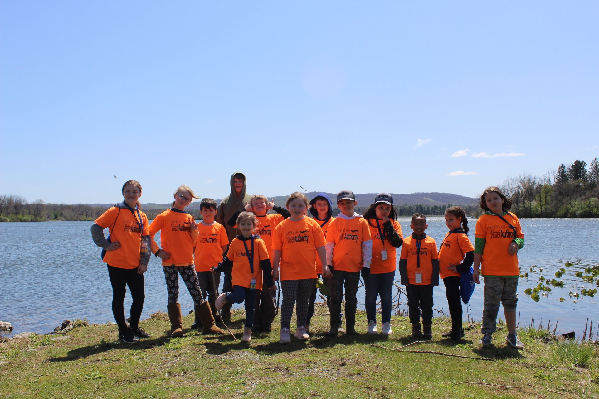 Group of children in orange shirts stand by a lake on a sunny day.