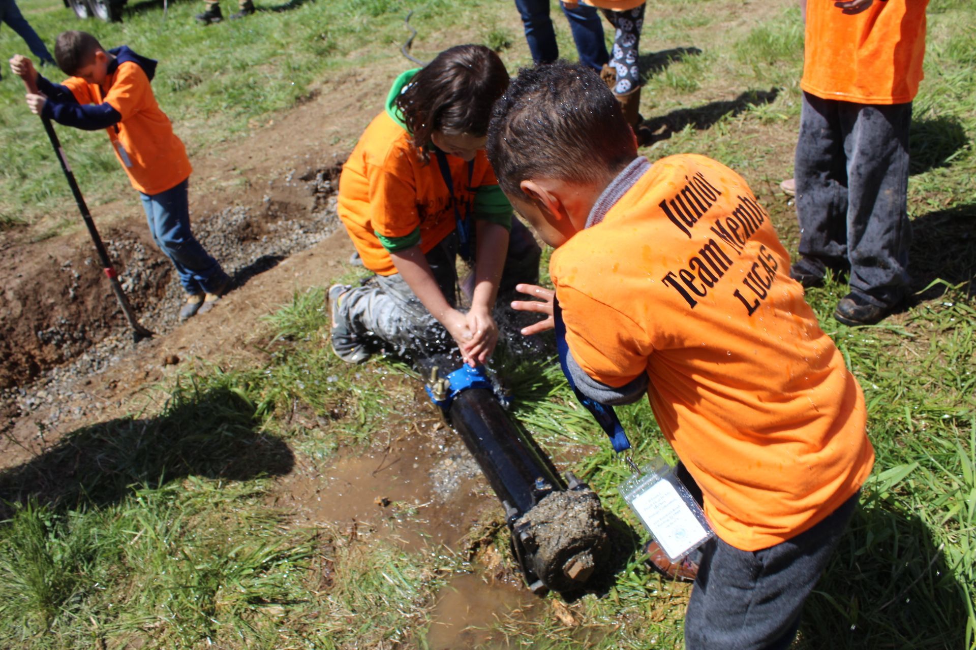 Students in orange shirts working on a pipe in a grassy field.