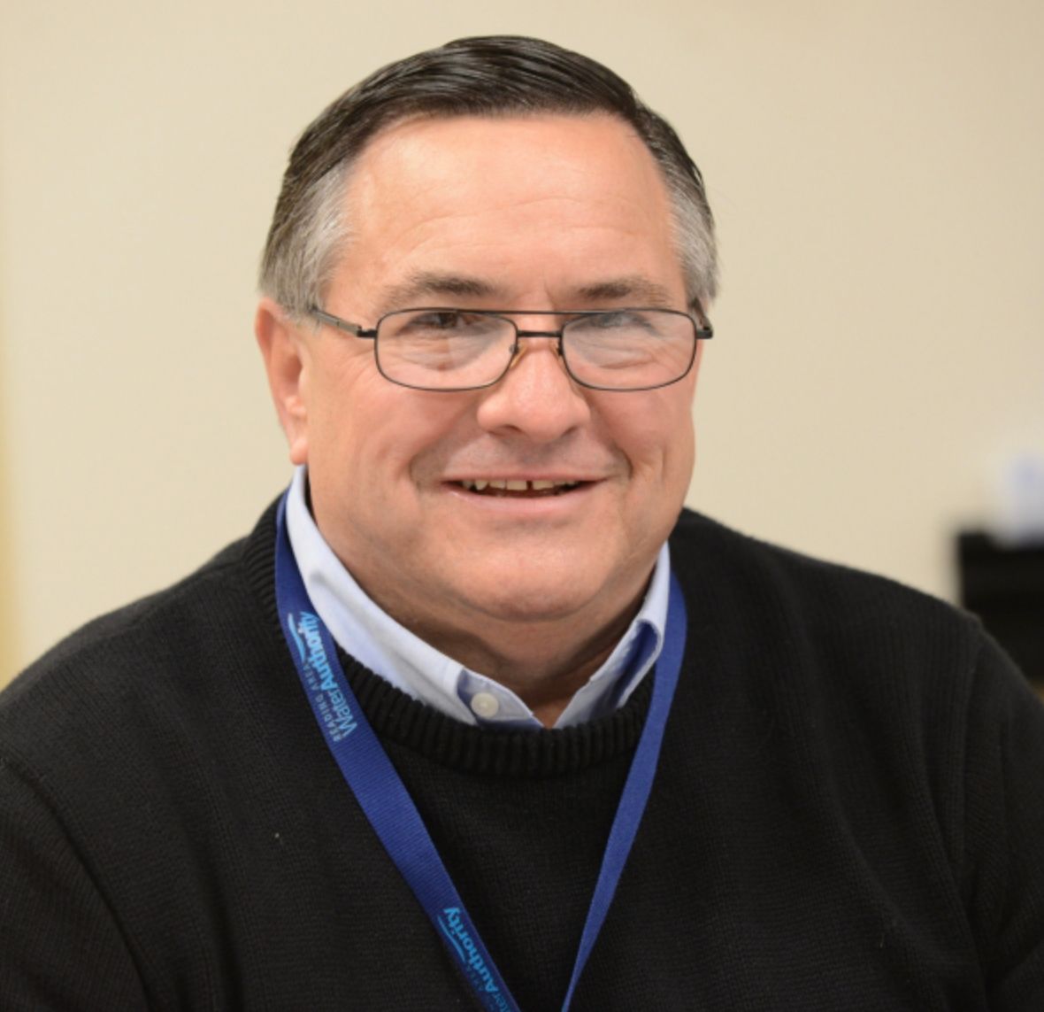 Man with glasses, smiling, wearing a blue lanyard, black sweater over a blue collared shirt.