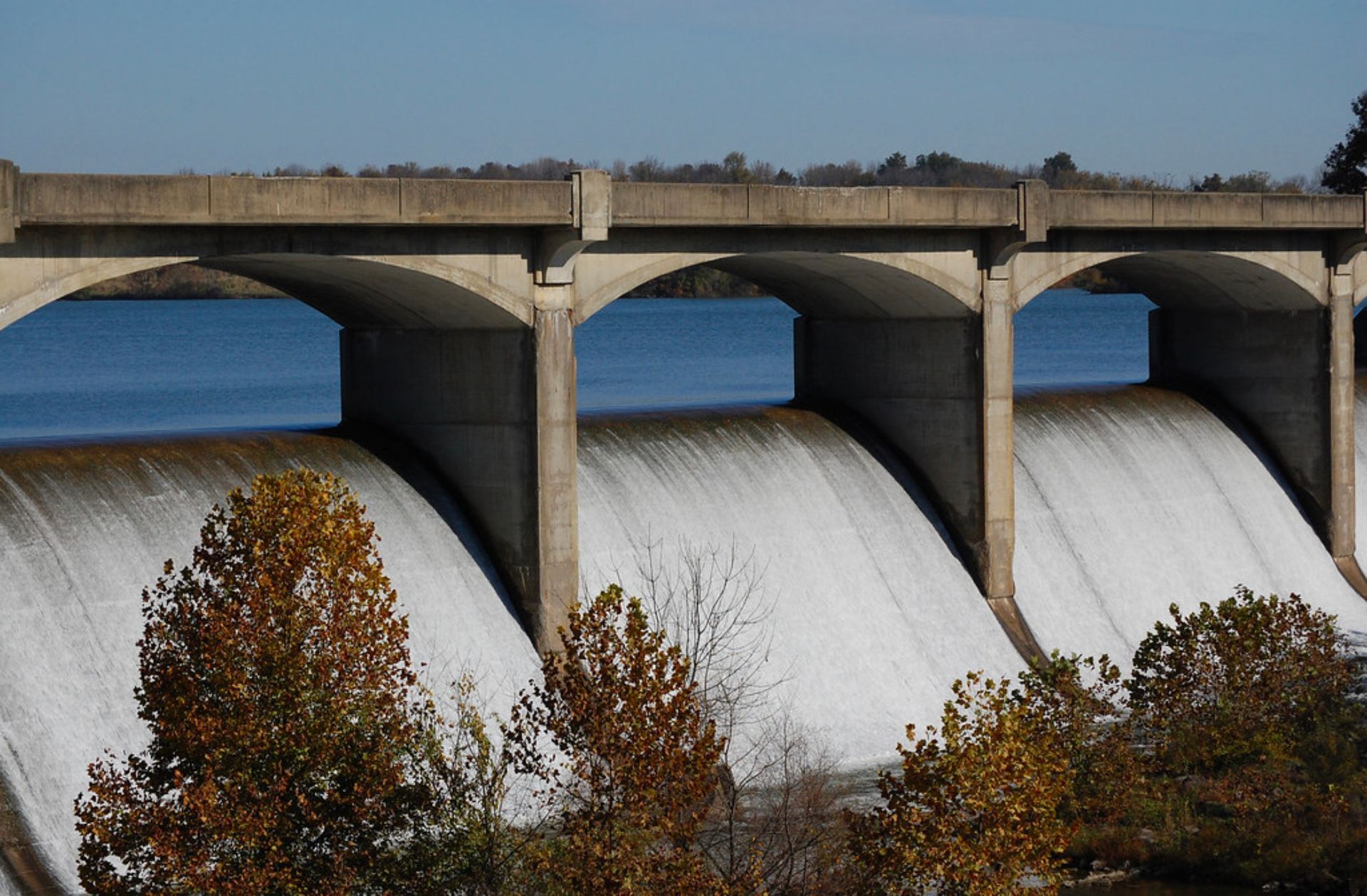 Dam with water cascading over its curved spillways; concrete structure; trees in foreground.