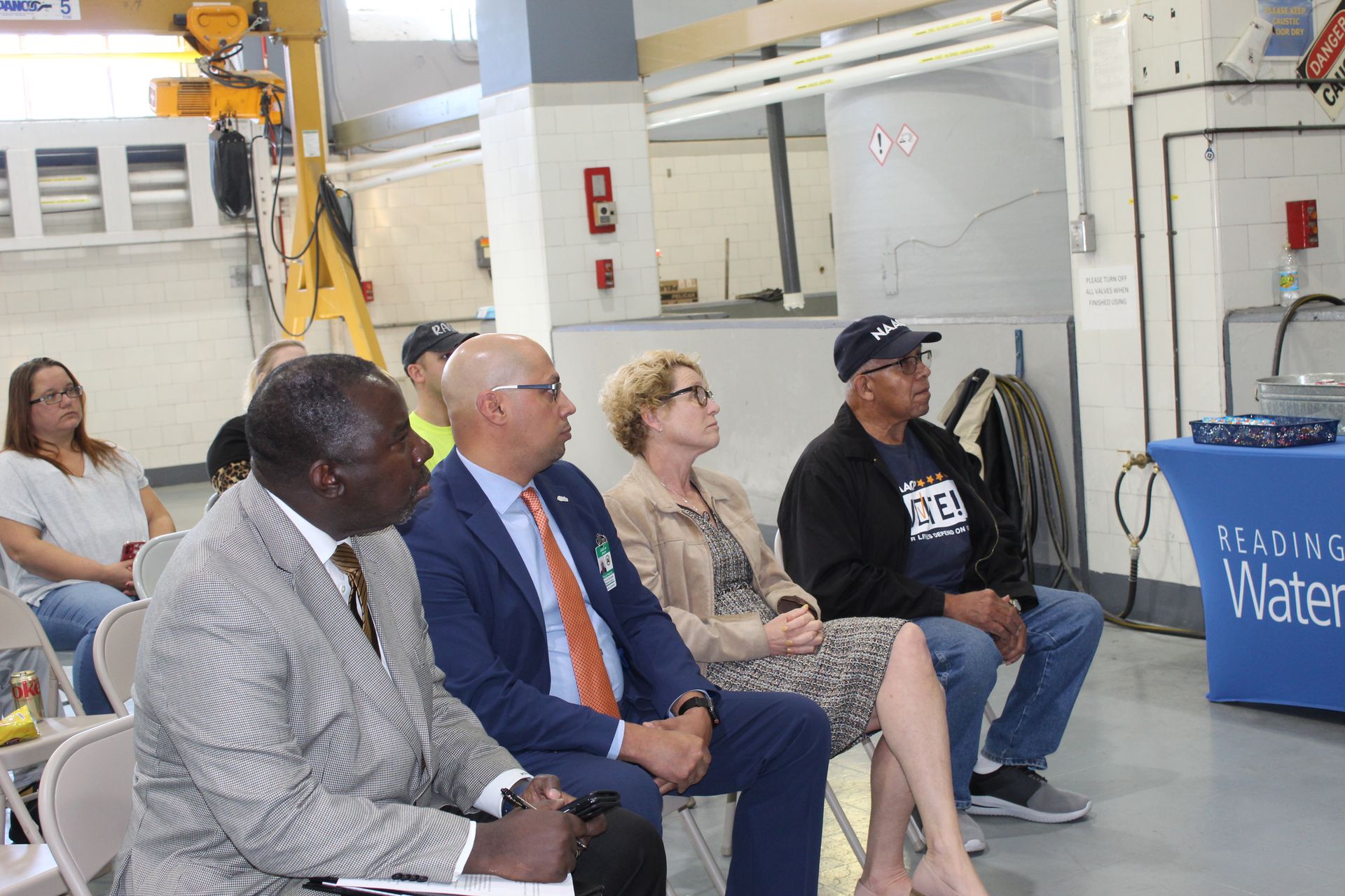 People seated, attending a presentation in a facility. Sign reads