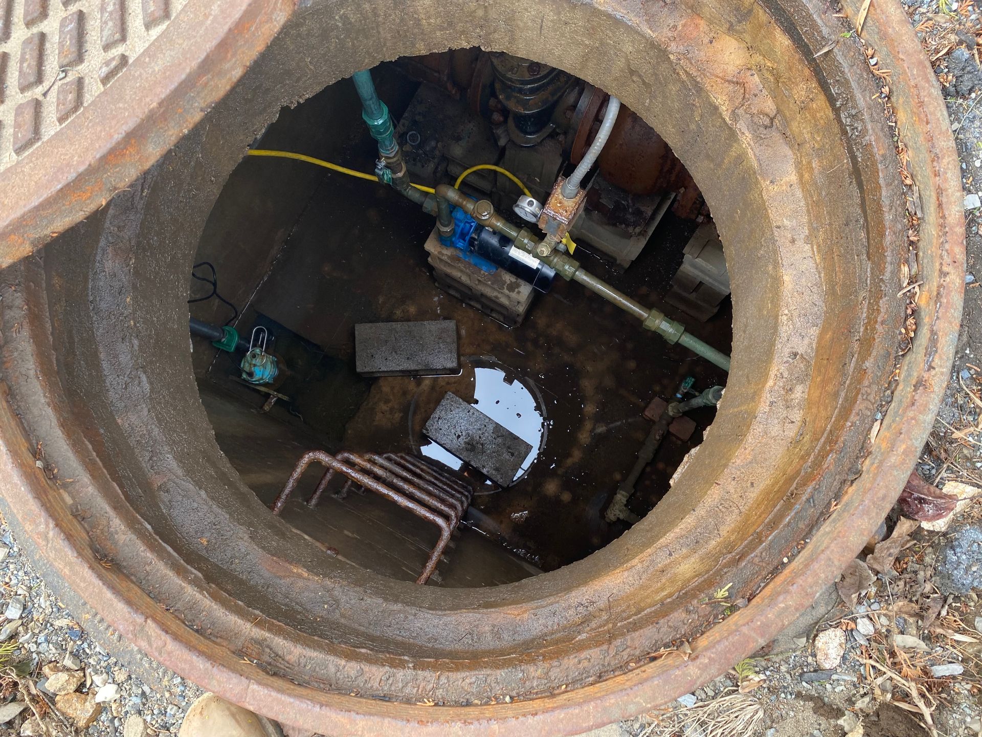 Open manhole showing pipes, a small ladder, and standing water. Dark interior.