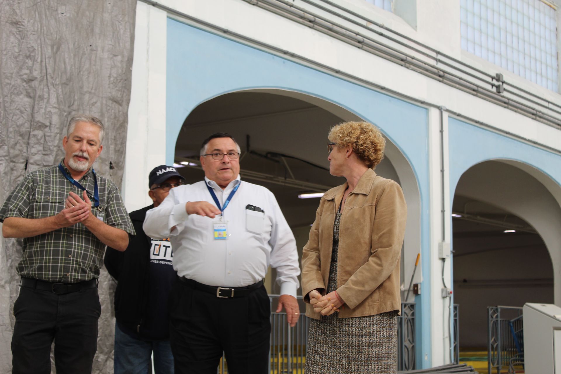 Three people indoors: One man gestures, others listen. Light blue arches in background.