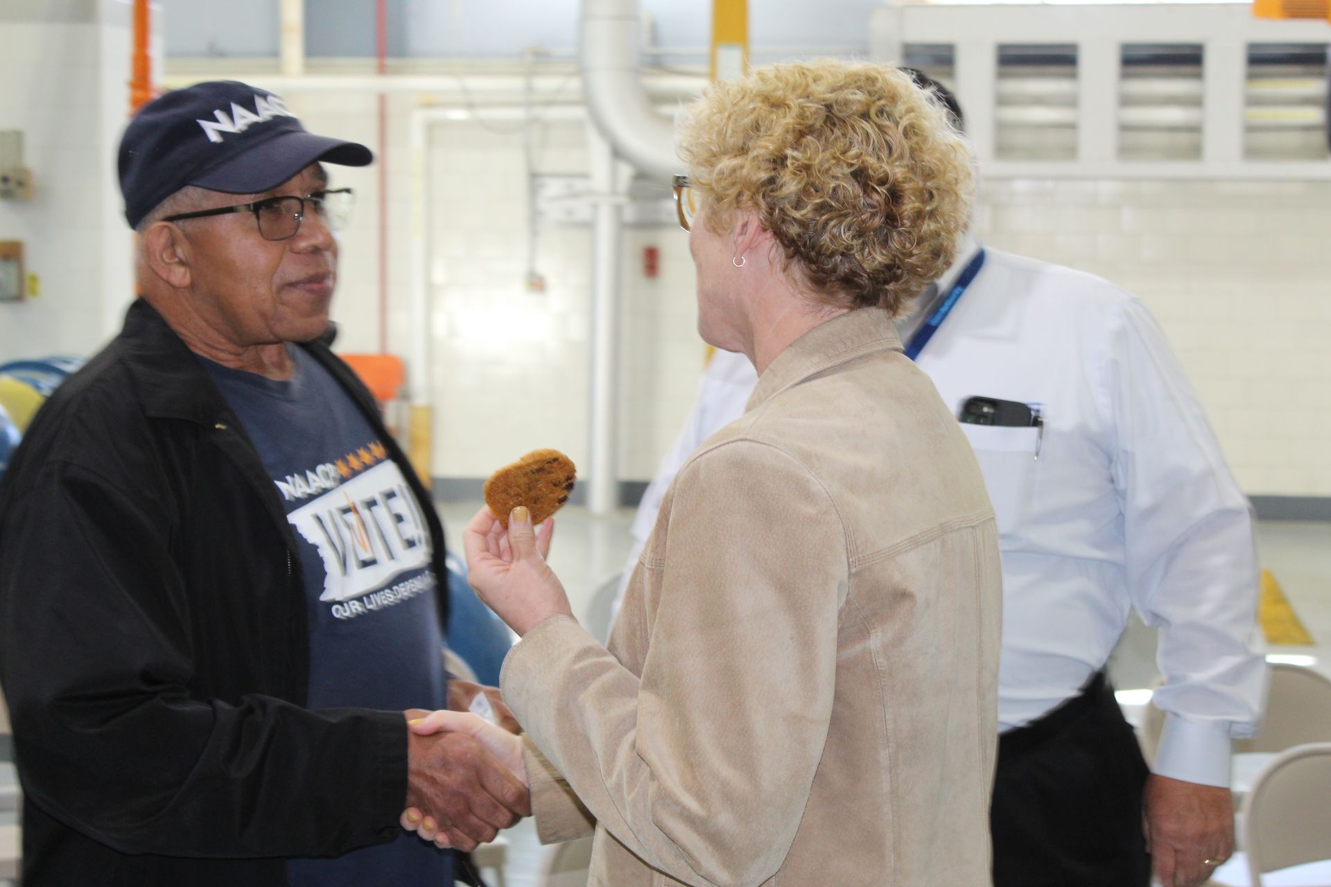 Man in a cap shakes hands with woman holding a pastry. Another person stands nearby. Indoors, industrial setting.