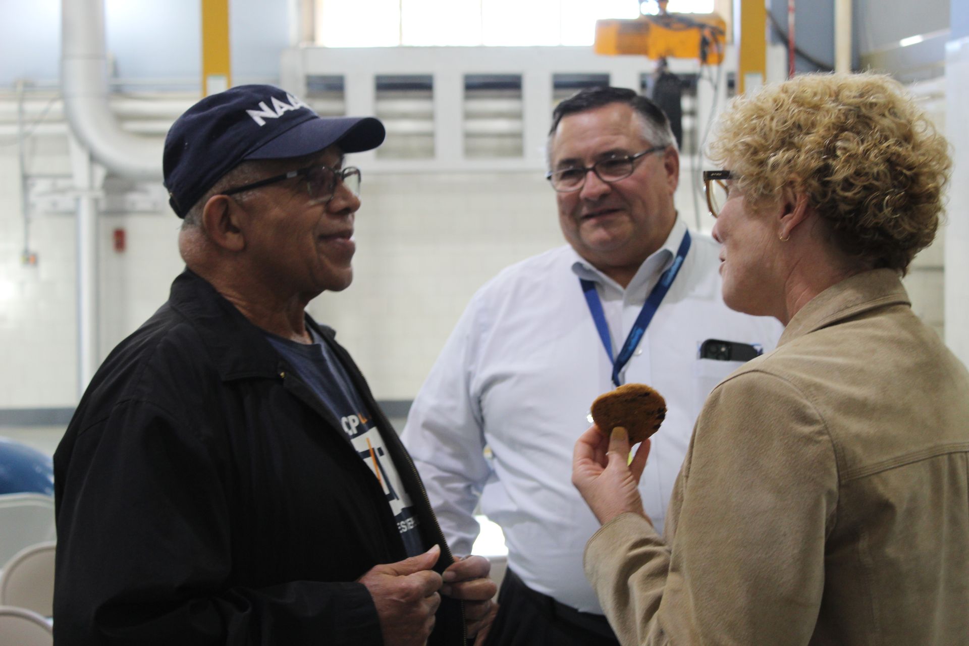 Three people conversing in a facility: man in cap, man in white shirt, woman holding cookie.