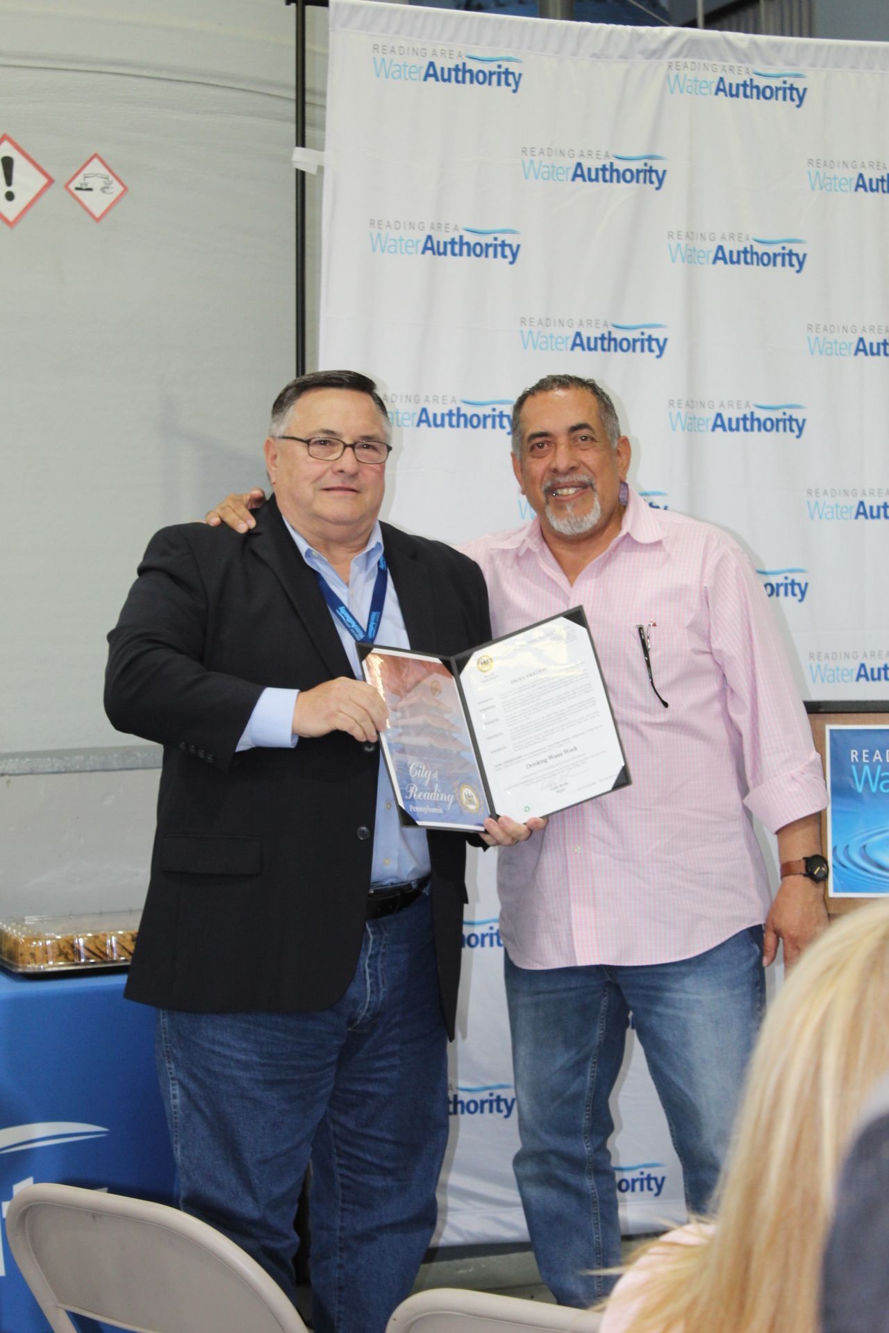 Two men holding an award and document near a blue backdrop. One man in a suit, the other in a pink shirt.